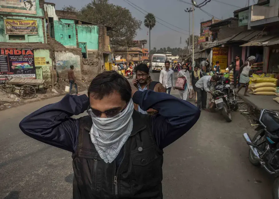 The open burning of trash  adds to the daily cloud. Here, a man ties a scarf over his face after encountering a smoldering pile of trash. Image by Larry C. Price. India, 2018.