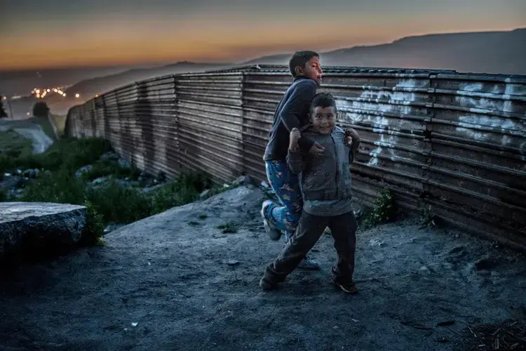 Two boys from the Arias family roughhouse in the dust beside their home abutting the original U.S.-built border wall in Colonia Libertad, Tijuana, Mexico. Behind them, a string of lights set up by U.S. Border Patrol stretches to the sea. The lights were set up by U.S. Border Patrol to illuminate the wall to make it more difficult migrants to cross at night. Image by James Whitlow Delano. Mexico, 2017.