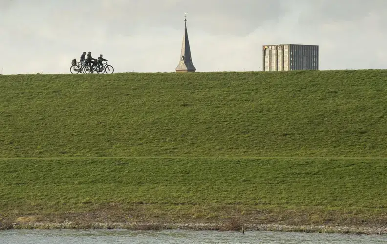 Cyclists ride by buildings peeking over a relocated levee in the town of Lent in the southeast corner of the Netherlands. The levee was moved to ease flood pressure on Nijmegen, an old river port across the Waal River from Lent. Image by Chris Granger. Netherlands, 2019.