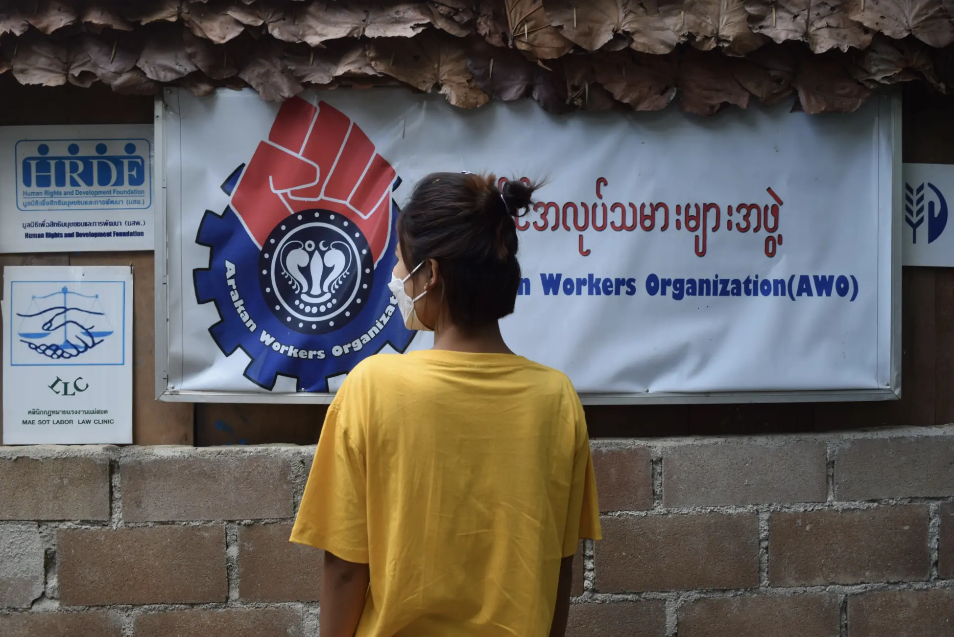 A woman in a yellow tee shirt stands masked in front of a sign that reads AWO Arakan Workers Organization.