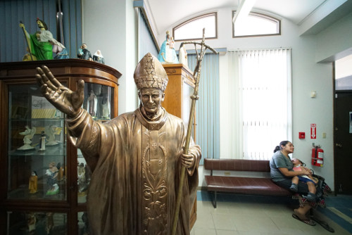 A woman waits with a child behind a statue in the Santa Barbara Catholic Church office in Dededo. Image by Cory Lum. Guam, 2017. A woman waits with a child behind a statue in the Santa Barbara Catholic Church office in Dededo. Image by Cory Lum. Guam, 2017.
