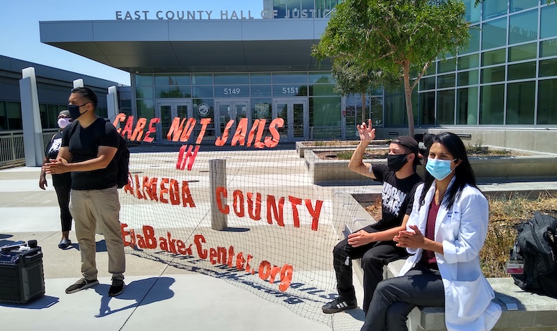 Three protestors stand outside Santa Rita Jail in front of a sign that says 'Care not jails in Alameda County - ellabakercenter.org.' Image by Andrew Stelzer / KALW. United States, undated. Three protestors stand outside Santa Rita Jail in front of a sign that says 'Care not jails in Alameda County - ellabakercenter.org.' Image by Andrew Stelzer / KALW. United States, undated.