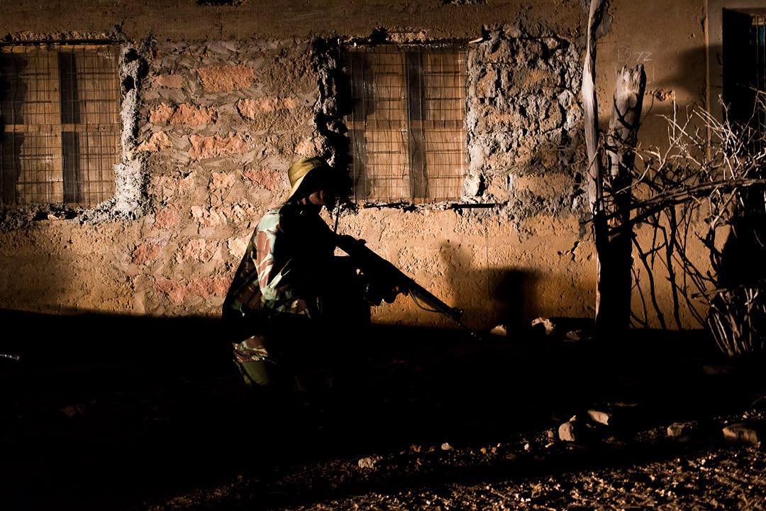 A Kenyan paramilitary policeman crouches in front of shuttered homes in Kenya’s northeast town of Mandera on the border with Somalia. All along this border, security is being bolstered by the Kenya Police Reserves, a locally recruited paramilitary force that speaks Somali and knows the community. Now that school is back in session, Roughly 300 Kenyan Somali policemen are stationed at nearby Garissa University in order to provide security services to the recently wounded area. Image by Will Swanson. Kenya, 2015. Media file: kenyaparamilitary.jpg