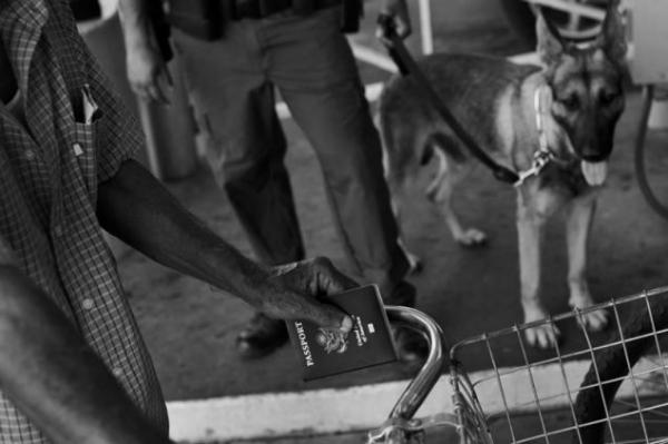 A man holding a U.S. passport coming from Mexico with his bike lines up at port of entry into the United States under the watchful eye of a U.S. Border Patrol Agent and a dog used to sniff out illegal narcotics in Laredo, Texas. Image by © Louie Palu/ZUMA. United States, 2012. Media file: louie2.jpg