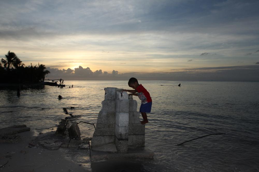 Four-year-old Aberam Maerere plays on the concrete block corner of a house that was claimed by the sea. The community of Te Bikenikora has lost dozens of homes in recent years to erosion and the advancing sea. The local church has elevated its foundation by 18 inches so parishioners could stay dry at high tide. Image by Kenneth R. Weiss. Kiribati, 2014. Media file: img_4753.jpg