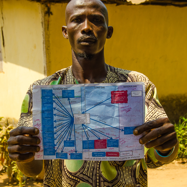 A quarantined man points out his position on a diagram that charts all of the contacts stemming from a single case of Ebola. Image by Amy Maxmen. Sierra Leone, 2014. Media file: _dsc9031.jpg