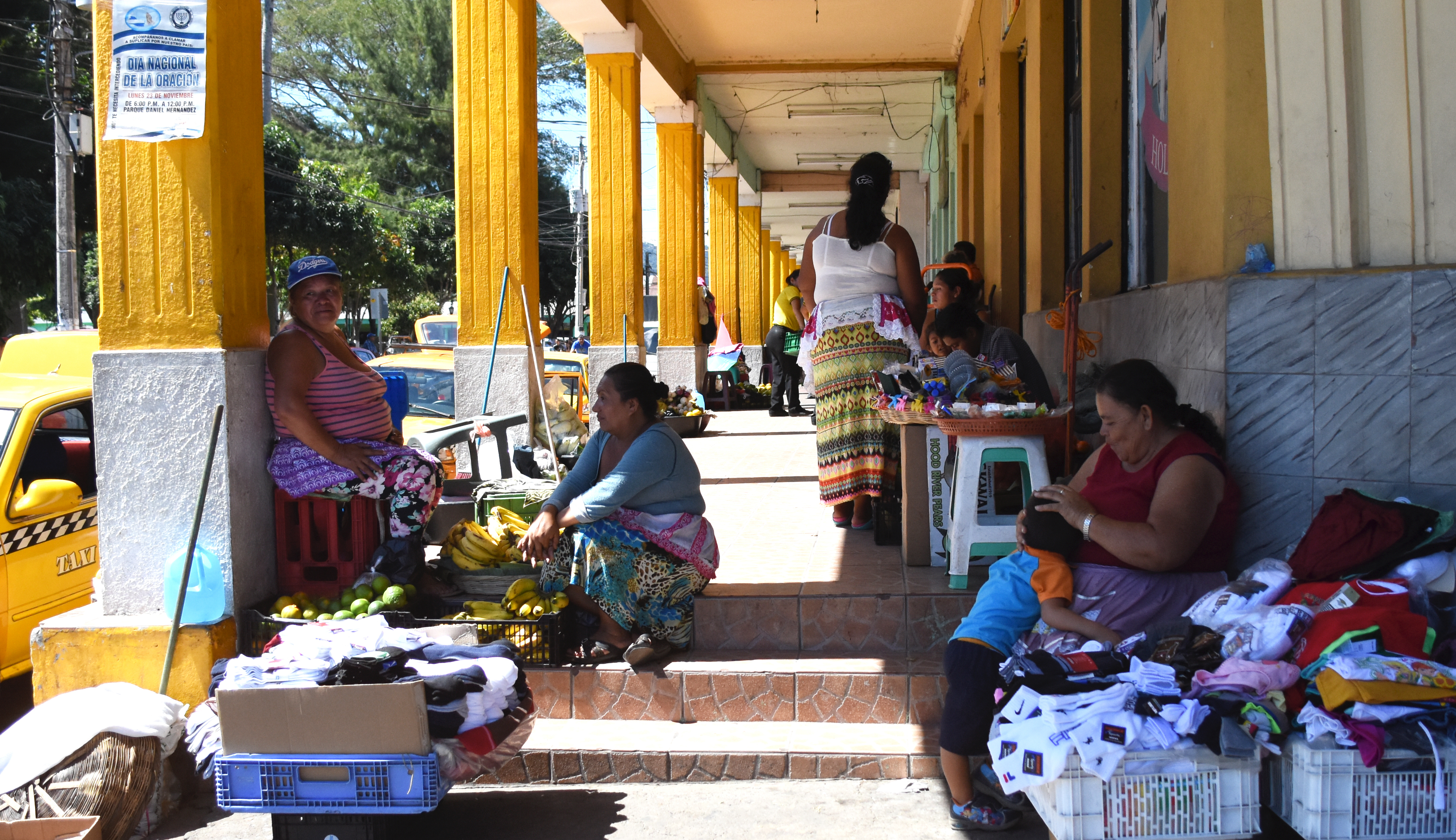Women and children sell fruit and bundles of socks on the sidewalk in Santa Ana, El Salvador. With minimum wage at just above $1, many people take to the streets selling small items in hopes to turn a profit. Children of all ages accompany their guardians in the street markets. Image by Kayli Plotner. El Salvador, 2016. Media file: santaana.jpg