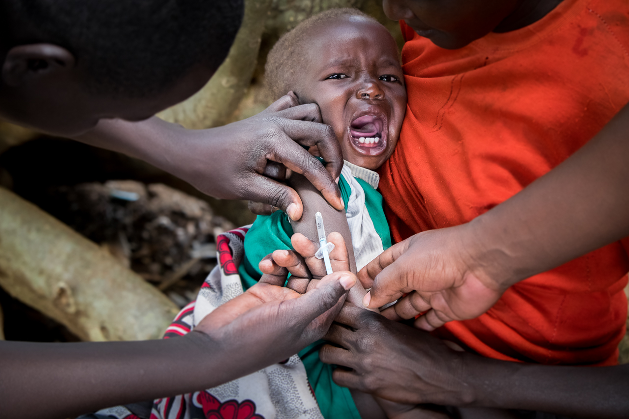A child receives an immunization as part of a Kenya Red Cross outreach program in rural West Pokot County, Kenya. For many in the area, the monthly mobile clinic, on a mat rolled out under a tree, is their only contact with the health system. Image by Paul Nevin. Kenya, 2014. Media file: 8.jpg