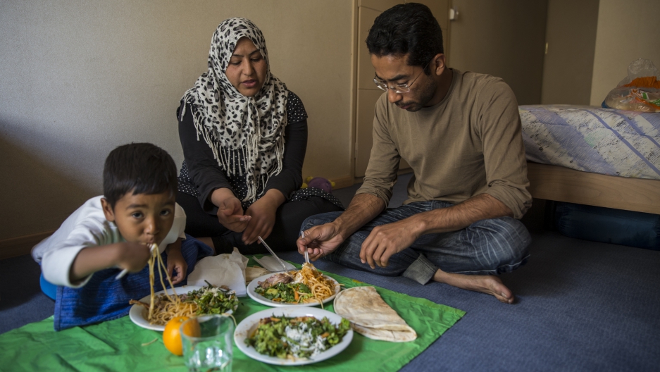 Ali Jaffari and his wife, Wajiha, and son Shayan, 4, eat dinner in their room while their other son naps. Together they traveled from Afghanistan to Greece, hoping to make their way to Germany. Now they are stuck in Greece. Image by Jodi Hilton. Greece, 2016. Media file: 050816_theworld_cityplaza38.jpg