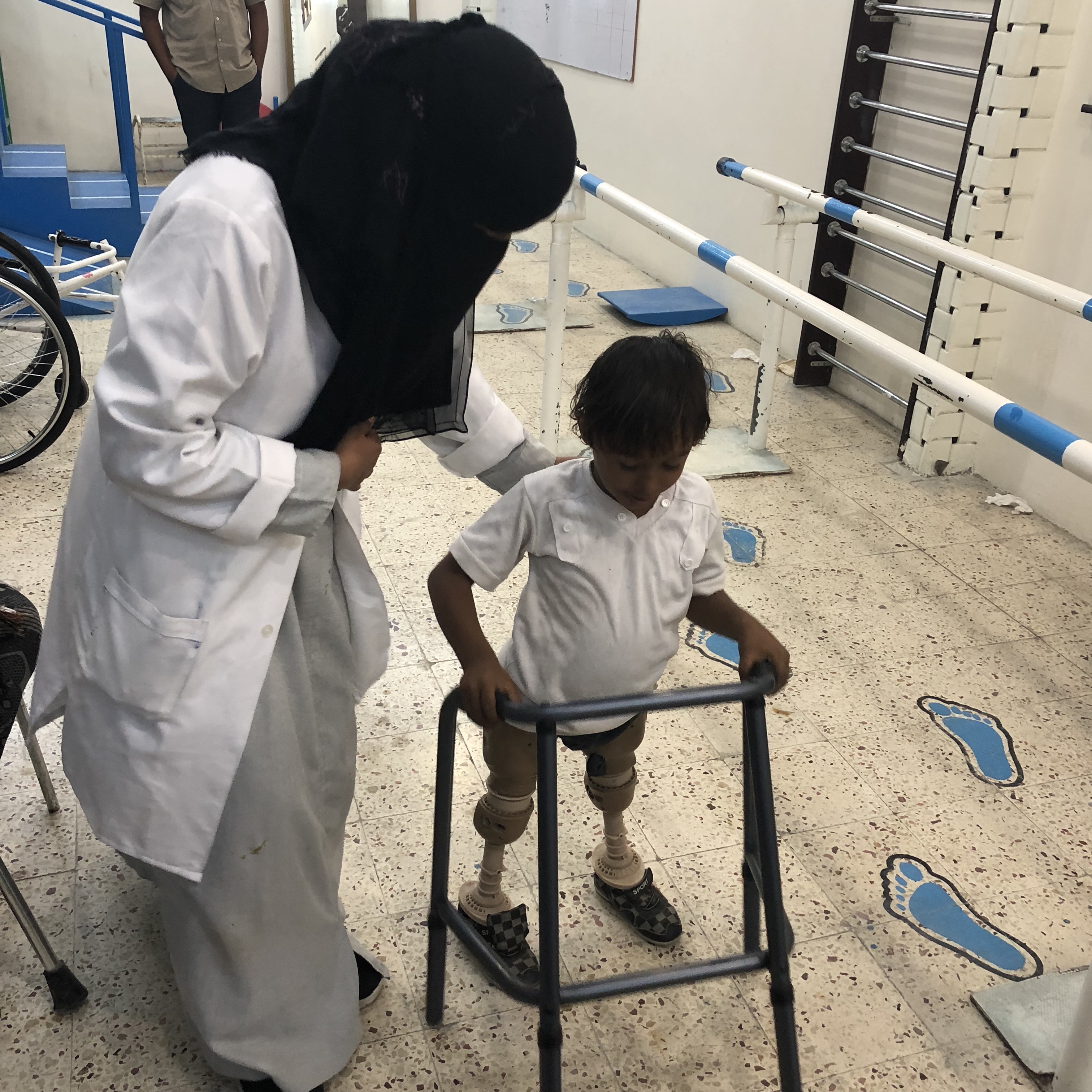 8 year old Imad lost his legs when his home was bombed in 2015. Samah Mohamed, his prosthetics technician, tries to get him to wear these prosthetics, but they are old, heavy, and don’t bend at the knee. Instead he prefers to scoot around on his hands. He’s one of many children with amputations being treated at the Prosthetic and Physiotherapy Center in Aden. Screenshot from PBS NewsHour. 8 year old Imad lost his legs when his home was bombed in 2015. Samah Mohamed, his prosthetics technician, tries to get him to wear these prosthetics, but they are old, heavy, and don’t bend at the knee. Instead he prefers to scoot around on his hands. He’s one of many children with amputations being treated at the Prosthetic and Physiotherapy Center in Aden. Screenshot from PBS NewsHour.