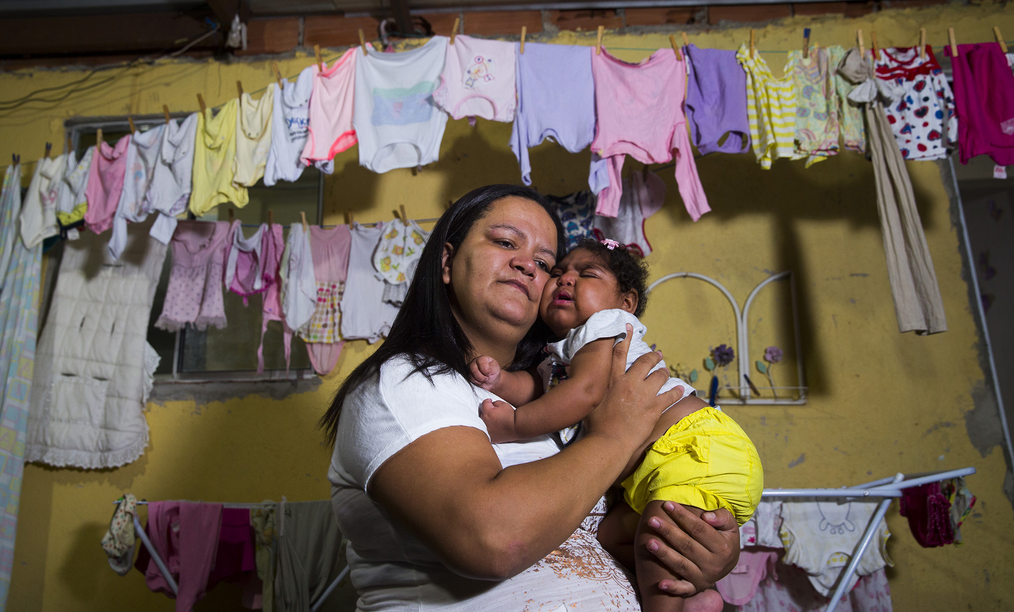 Fabiane Lopes holds her daughter, Valentina at their home in Duque de Caxias, a suburb of Rio de Janeiro in southeastern Brazil. Valentina's mother was pregnant when she became infected with the Zika virus and her daughter was born with microcephaly—a congenital malformation with smaller than normal head size for age and sex as well as other profound birth defects. Brazil has confirmed far more malformations of the brain in babies born to mothers who were infected with Zika than any other country. After she gave birth to Valentina, she elected to have a tubal ligation so she could not become pregnant again. Image by Mark Hoffman. Brazil, 2017. Fabiane Lopes holds her daughter, Valentina at their home in Duque de Caxias, a suburb of Rio de Janeiro in southeastern Brazil. Valentina's mother was pregnant when she became infected with the Zika virus and her daughter was born with microcephaly—a congenital malformation with smaller than normal head size for age and sex as well as other profound birth defects. Brazil has confirmed far more malformations of the brain in babies born to mothers who were infected with Zika than any other country. After…