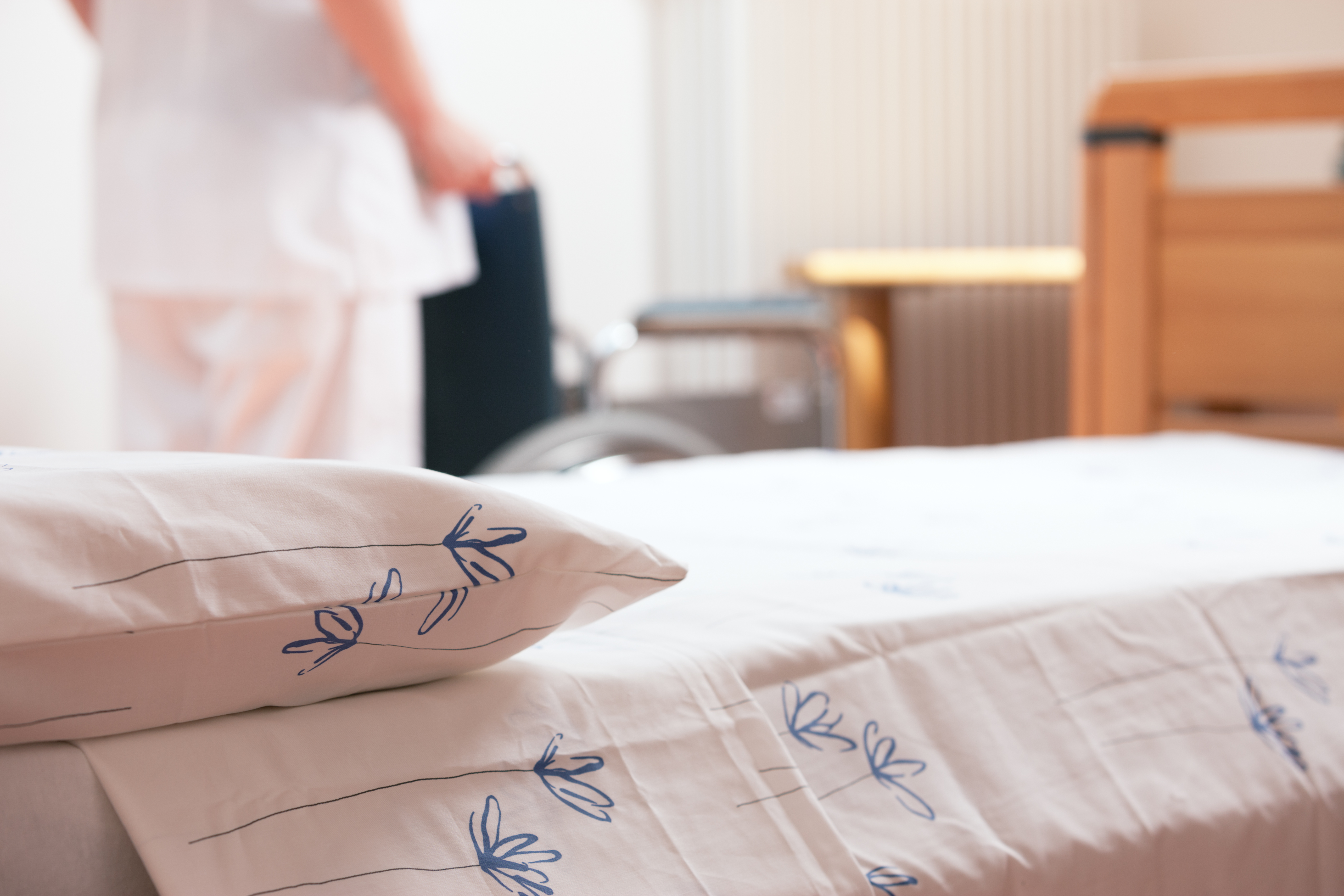 Bed at nursing home with nurse and wheelchair in background. Image by Stokkete / Shutterstock.com. Undated. Bed at nursing home with nurse and wheelchair in background. Image by Stokkete / Shutterstock.com. Undated.