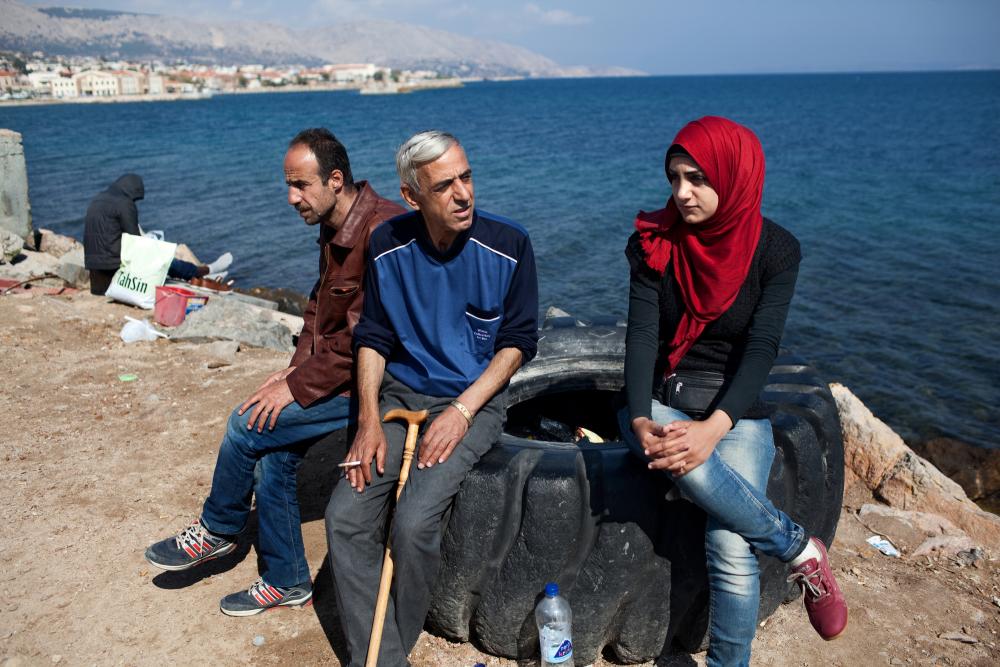 Left to right, Muhannad, Ali, and Ali's daughter Salam, all from Yarmouk Palestinian camp near Damascus, idly pass the time at the Coast Guard detention camp on Chios Island, Greece. Migrants who are found or rescued nearby spend at least a couple of days here before going to the police detention camp. Image by Holly Pickett. Greece, 2014.