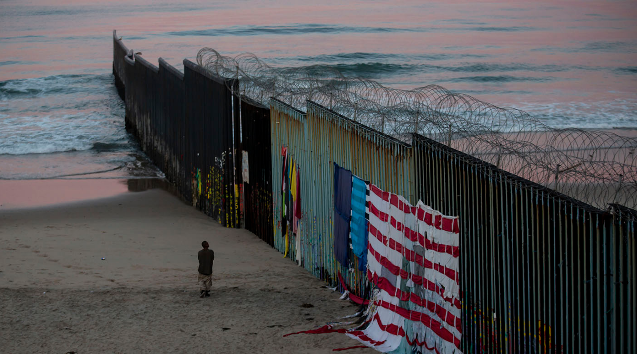 In Tijuana, a man walks at sunrise along the border fence where it meets the Pacific Ocean. Image by Erika Schultz. Mexico, 2019. In Tijuana, a man walks at sunrise along the border fence where it meets the Pacific Ocean. Image by Erika Schultz. Mexico, 2019.