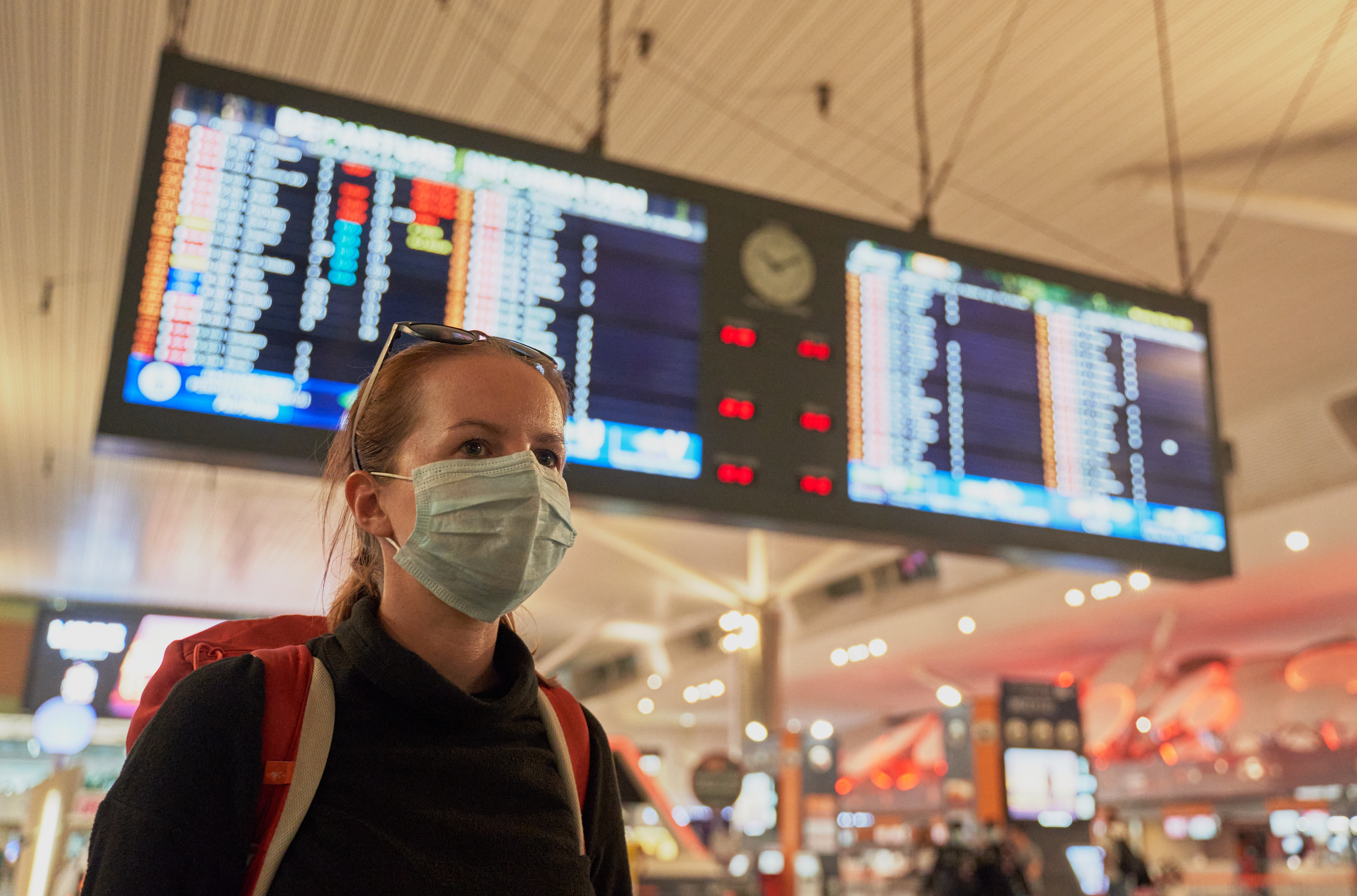 A woman with a backpack wears an anti-virus mask on her face at an airport. Image by Damian Pankowiec/Shutterstock. 2020. A woman with a backpack wearing an anti-virus mask on her face at the airport arrivals board. Image by Damian Pankowiec/Shutterstock. 2020.
