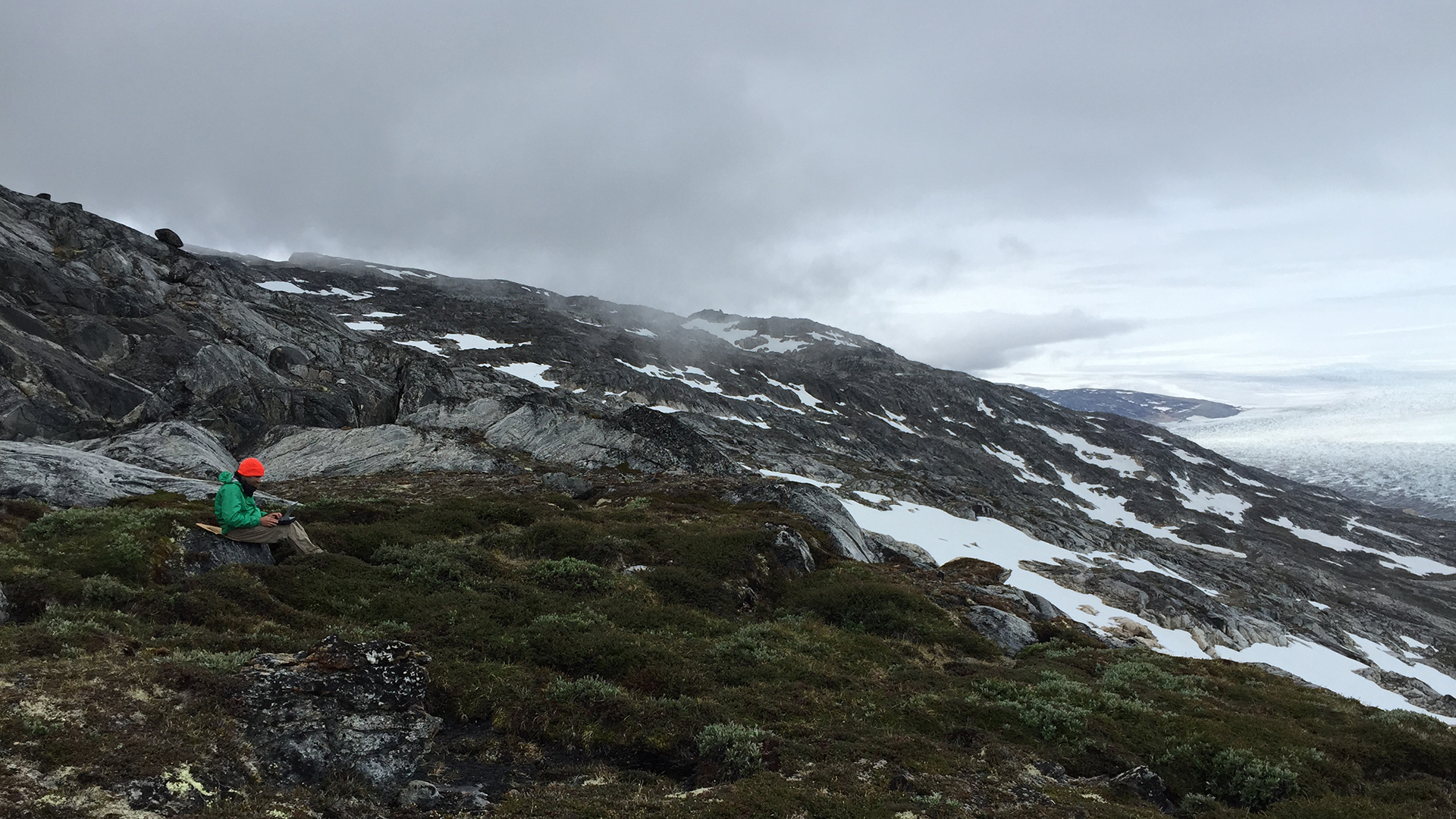 Science reporter Ari Daniel on assignment in Greenland. Helheim glacier, one of Greenland's largest glaciers, is off to the right. Image by Kate Raisz. Greenland, 2015. Media file: ariorange.jpg