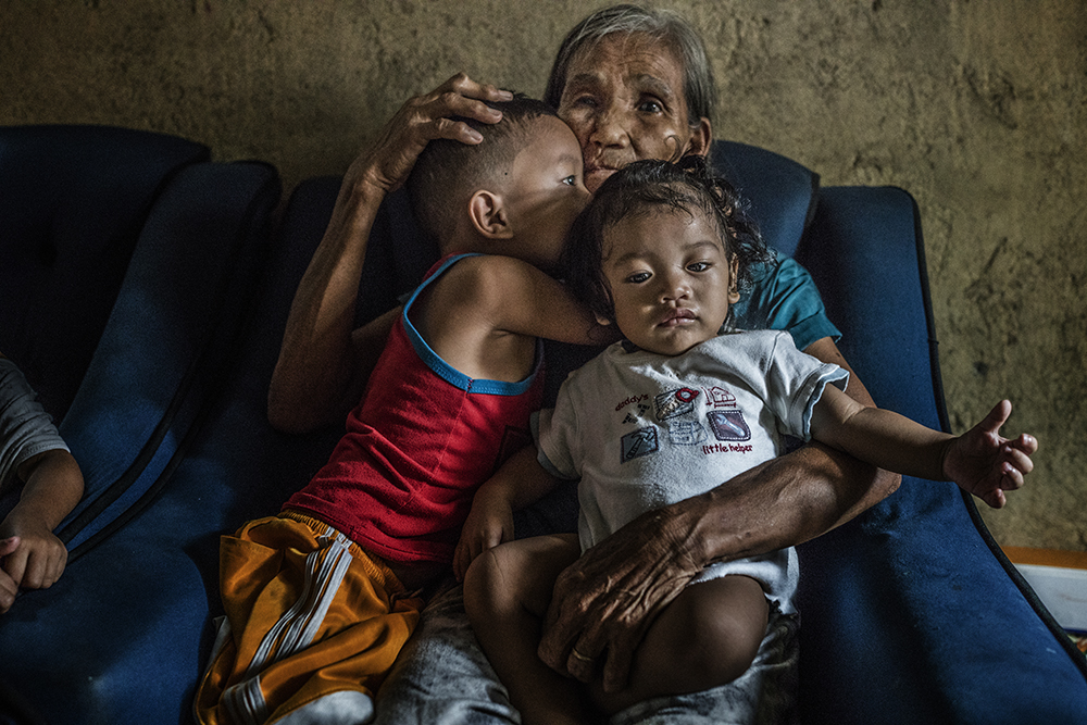 Duterte’s war on drugs leaves tragic legacy for Filipino families Remy Fernandez, 84 years old, holds two of the seven grandchildren she is raising. Her youngest son, Constantino de Juan, a methamphetamine user, was killed by masked men in December, 2016. Upon seeing his attackers, Juan instructed five-year-old CJ, shown here wearing a red tank top, to take care of his siblings. The children's mother is in prison due to a drug arrest. Baby RJ, in the "daddy's little helper" T-shirt, was born in prison. Image by James Whitlow Delano. Philippines, 2017.