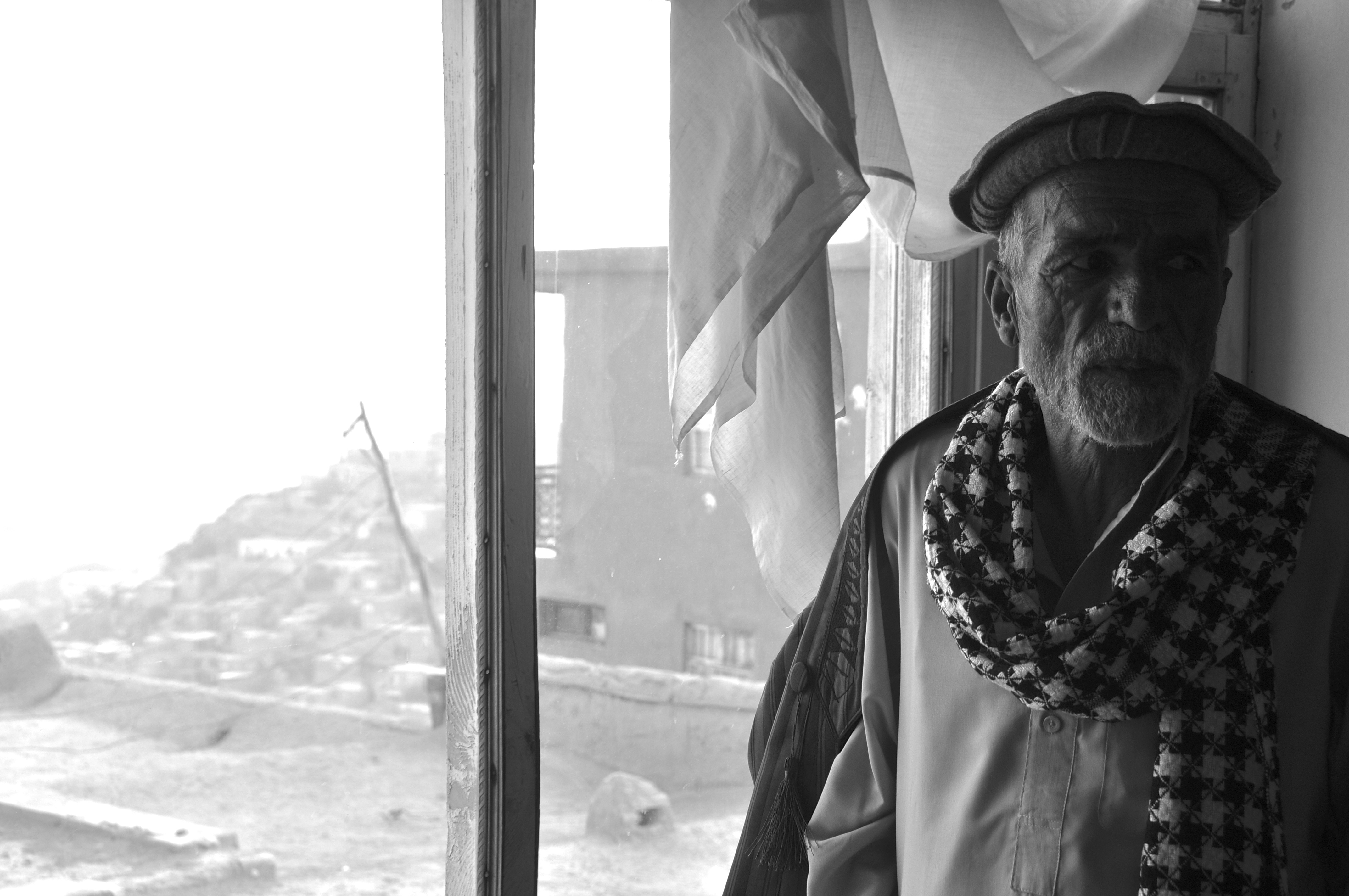 Wakil Abdul Rahim stands in front of a window in his mountaintop house, overlooking Kabul. Forty years ago, Rahim claimed land on the mountain by building on it. Back then, his was one of the only houses. Now there are tens of thousands of squatter houses on the mountains surrounding the city. Image by Jeffrey E. Stern. Afghanistan, 2013. Media file: dsc_0032.jpg