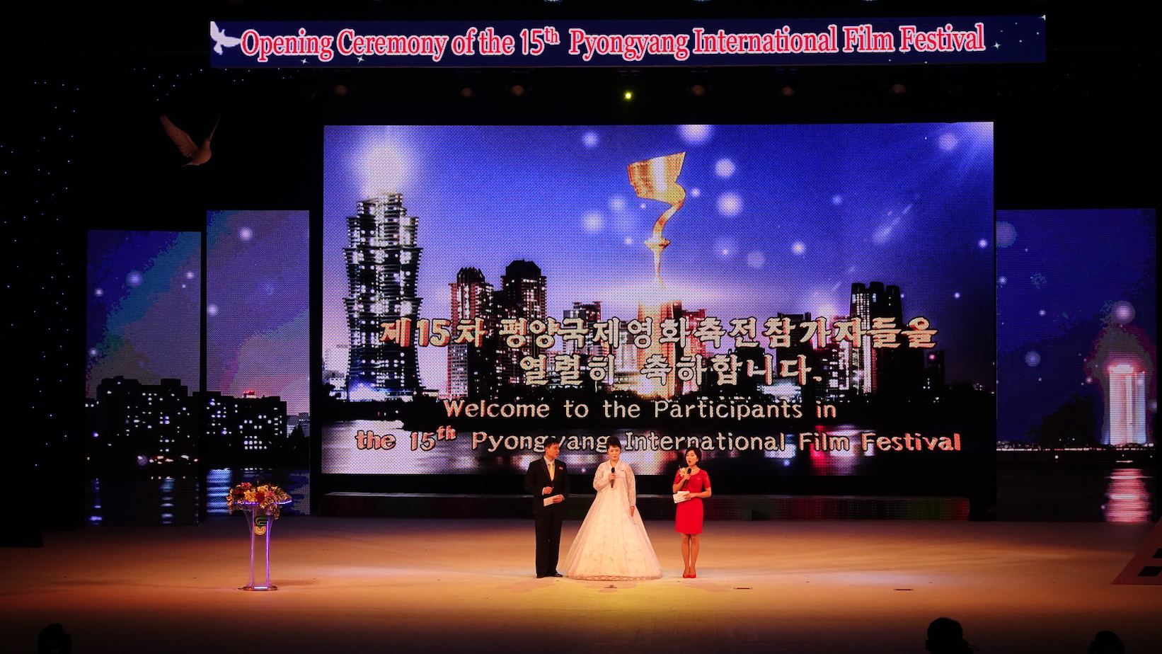 The three hosts on stage during the opening ceremony of the 15th Pyongyang International Film Festival. A sculpted dove in the top left signifies the festival's mission of peace. Image by Laya Maheshwari. North Korea, 2016. The three hosts on stage during the opening ceremony of the 15th Pyongyang International Film Festival. A sculpted dove in the top left signifies the festival's mission of peace. Image by Laya Maheshwari. North Korea, 2016.