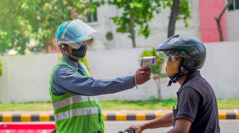 A medical worker in Jamshedpur checks the body temperature of a biker with an infrared thermometer gun for COVID-19 symptoms. Image by ArnavIG / Shutterstock. India, 2020. A medical worker in Jamshedpur checks the body temperature of a biker with an infrared thermometer gun for COVID-19 symptoms. Image by ArnavIG / Shutterstock. India, 2020.