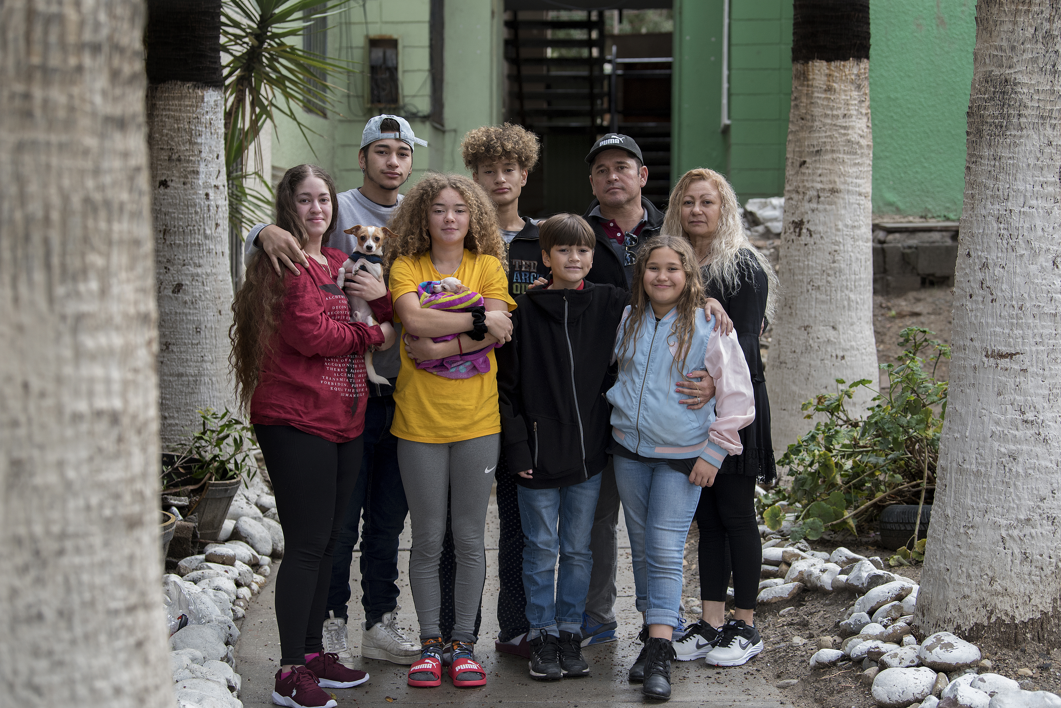 The Flores family pauses for a portrait outside Ramon’s apartment complex in Tijuana, Mexico. Family members are, clockwise from left, Leslie, 23, Lennes, 18, Raymond, 15, Ramon, 46, Enedis, 54, Rayma, 10, Edward, 12, and Kennedy, 16. The dogs are Canelo, left, and Bambi, wrapped in the blanket. Image by Amanda Cowan. Mexico, 2019. The Flores family pauses for a portrait outside Ramon’s apartment complex in Tijuana, Mexico. Family members are, clockwise from left, Leslie, 23, Lennes, 18, Raymond, 15, Ramon, 46, Enedis, 54, Rayma, 10, Edward, 12, and Kennedy, 16. The dogs are Canelo, left, and Bambi, wrapped in the blanket. Image by Amanda Cowan. Mexico, 2019.
