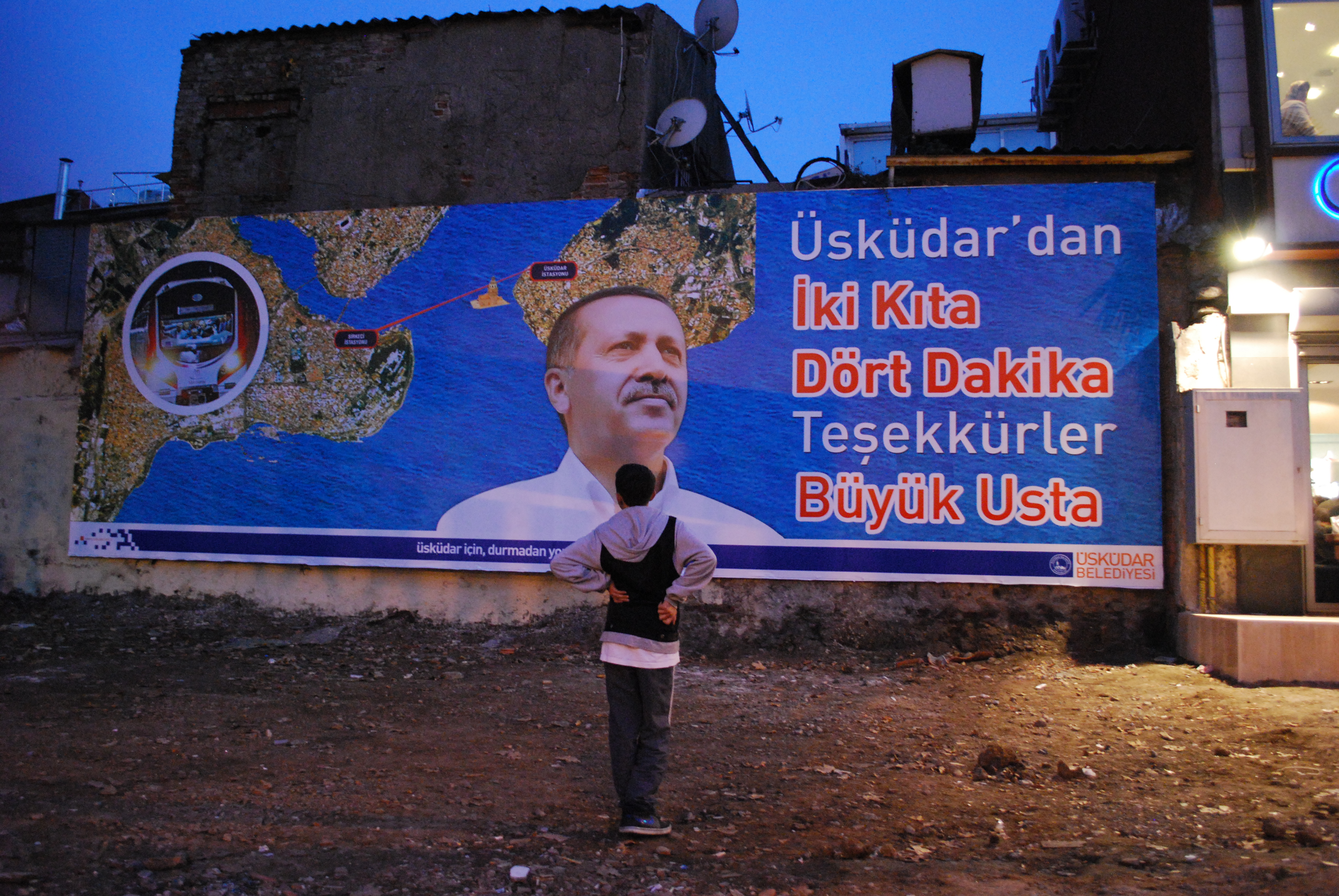 A kid gazes at a poster advertising the Marmaray project. The sign reads "Two Continents, Four Minutes," and thanks Erdogan, who is pictured. Image by Jenna Krajeski. Turkey, 2013. Media file: kidmarmaray.jpg