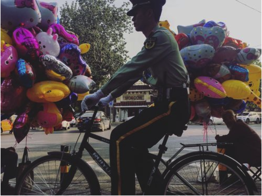 A member of the Chinese military rides his bike past two men selling balloons on Sunday, Sept. 24, 2017, on the same block as the residence of U.S. Ambassador to China Terry Branstad in Beijing, China. Image by Kelsey Kremer. China, 2017. A member of the Chinese military rides his bike past two men selling balloons on Sunday, Sept. 24, 2017, on the same block as the residence of U.S. Ambassador to China Terry Branstad in Beijing, China. Image by Kelsey Kremer. China, 2017.
