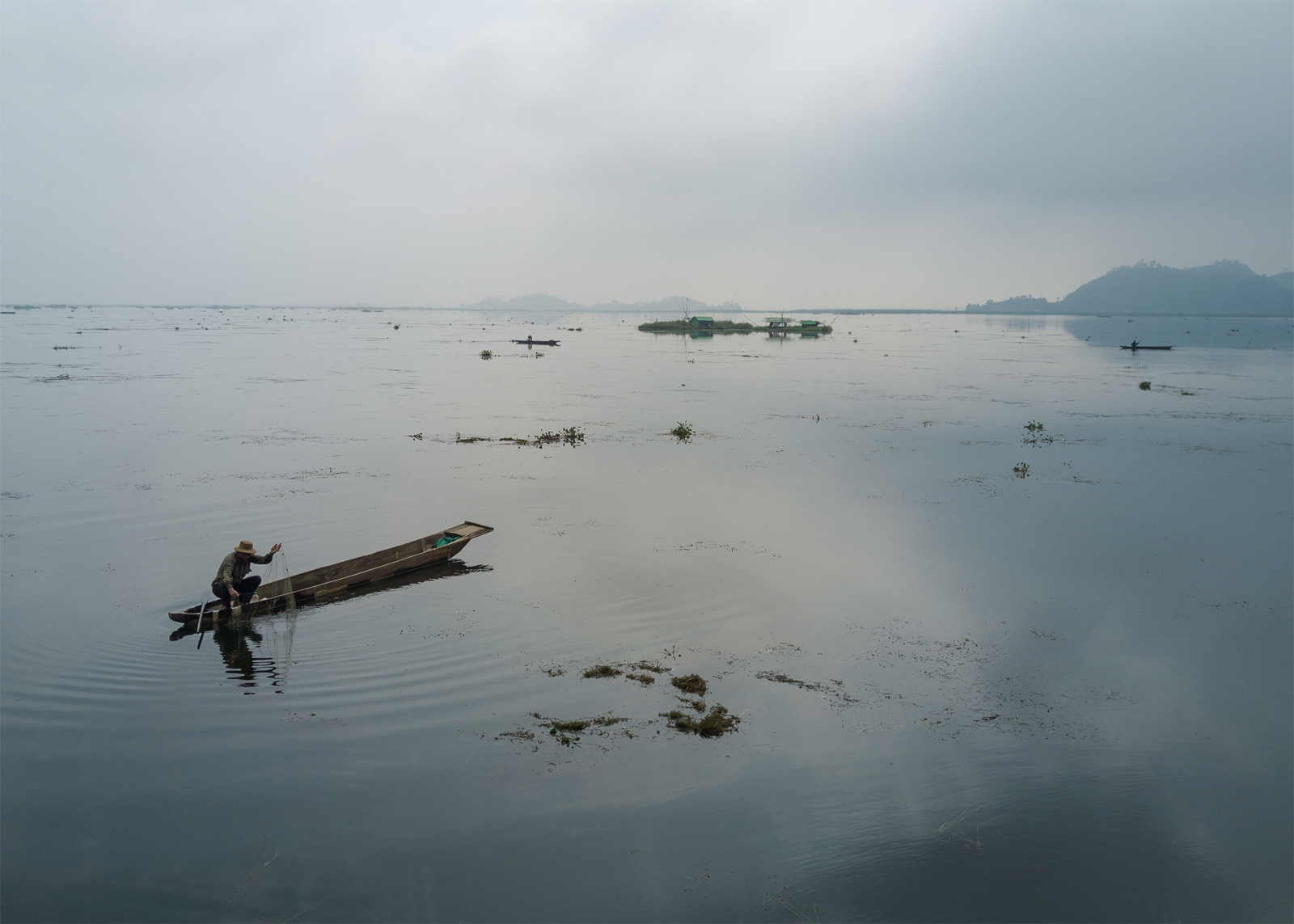 A Meitie fisherman fishes near phumshangs (floating huts) in Loktak Lake, Manipur. The construction of the Ithai Dam flooded over 80,000 hectares of farm and pasture land in the vicinity of the Loktak wetland. The Loktak Development Authority has been dredging the phumdis (floating biomass) and has cleared over 800 Phumshangs in the name of lake clean-up for efficient power generation. Image by Neeta Satam. India, 2017. A Meitie fisherman fishes near phumshangs (floating huts) in Loktak Lake, Manipur. The construction of the Ithai Dam flooded over 80,000 hectares of farm and pasture land in the vicinity of the Loktak wetland. The Loktak Development Authority has been dredging the phumdis (floating biomass) and has cleared over 800 Phumshangs in the name of lake clean-up for efficient power generation. Image by Neeta Satam. India, 2017.