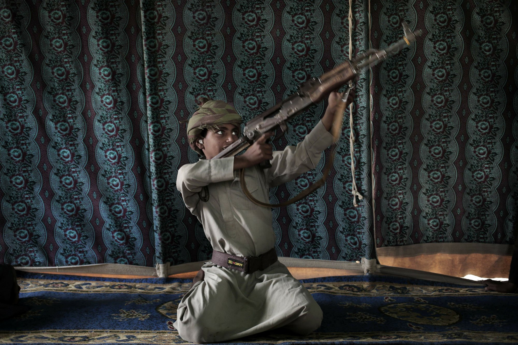 Kahlan, a 12-year-old former child soldier, demonstrates how to use a weapon, at a camp for displaced persons where he took shelter with his family, in Marib, Yemen in this July 27, 2018 photo. Houthi rebels took Kahlan and his classmates, promising to give them new school bags, but instead they were enlisted and trained as fighters tasked with carrying supplies to the front lines. There, he had to elude explosions and airstrikes that left mangled casualties on the battlefield. "The sight of the bodies was scary," he said. Image by Nariman El-Mofty / AP News. Yemen, 2018. Kahlan, a 12-year-old former child soldier, demonstrates how to use a weapon, at a camp for displaced persons where he took shelter with his family, in Marib, Yemen in this July 27, 2018 photo. Houthi rebels took Kahlan and his classmates, promising to give them new school bags, but instead they were enlisted and trained as fighters tasked with carrying supplies to the front lines. There, he had to elude explosions and airstrikes that left mangled casualties on the battlefield. "The sight of the bodies was…