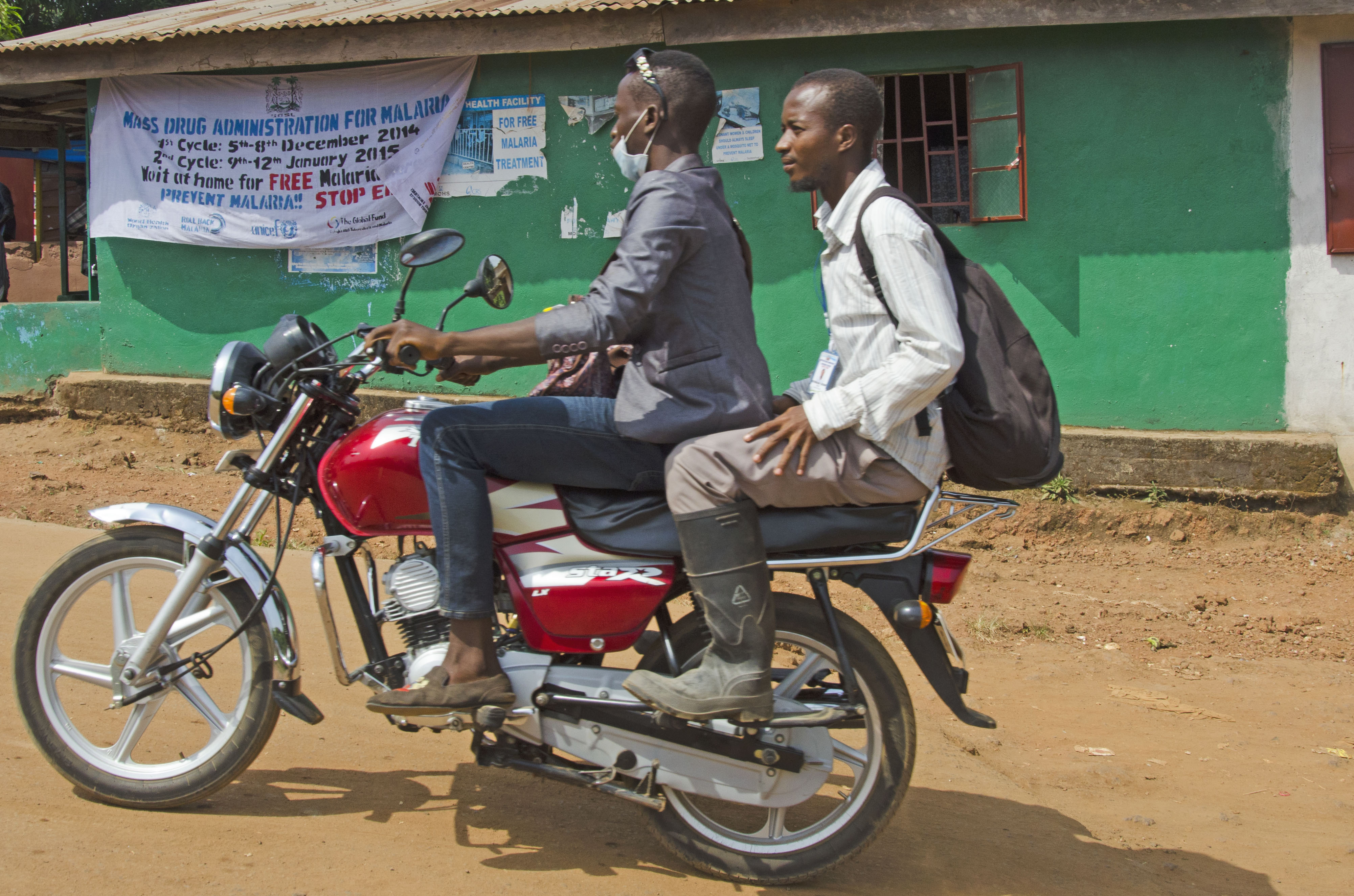 Two men on a motorbike pass a banner announcing a malaria prevention initiative in Sierra Leone. Image by Amy Maxmen. Sierra Leone, 2014.