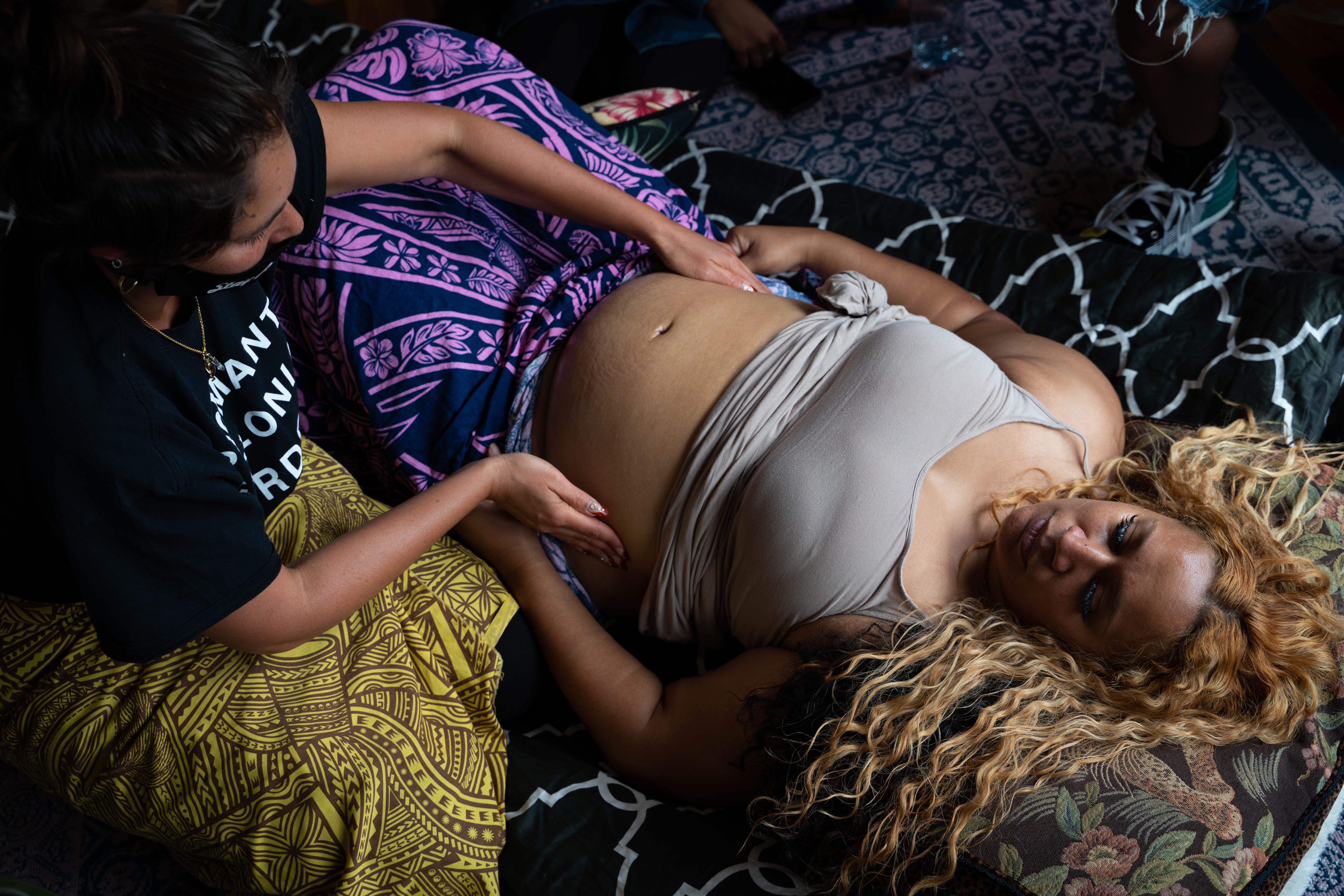 Overhead shot of a pregnant woman lying on a bed with her eyes closed, her head at the bottom right of the frame, and her stomach at the top left of the frame. Her tank top is rolled up, and someone to her left is placing their hands on either side of her stomach.