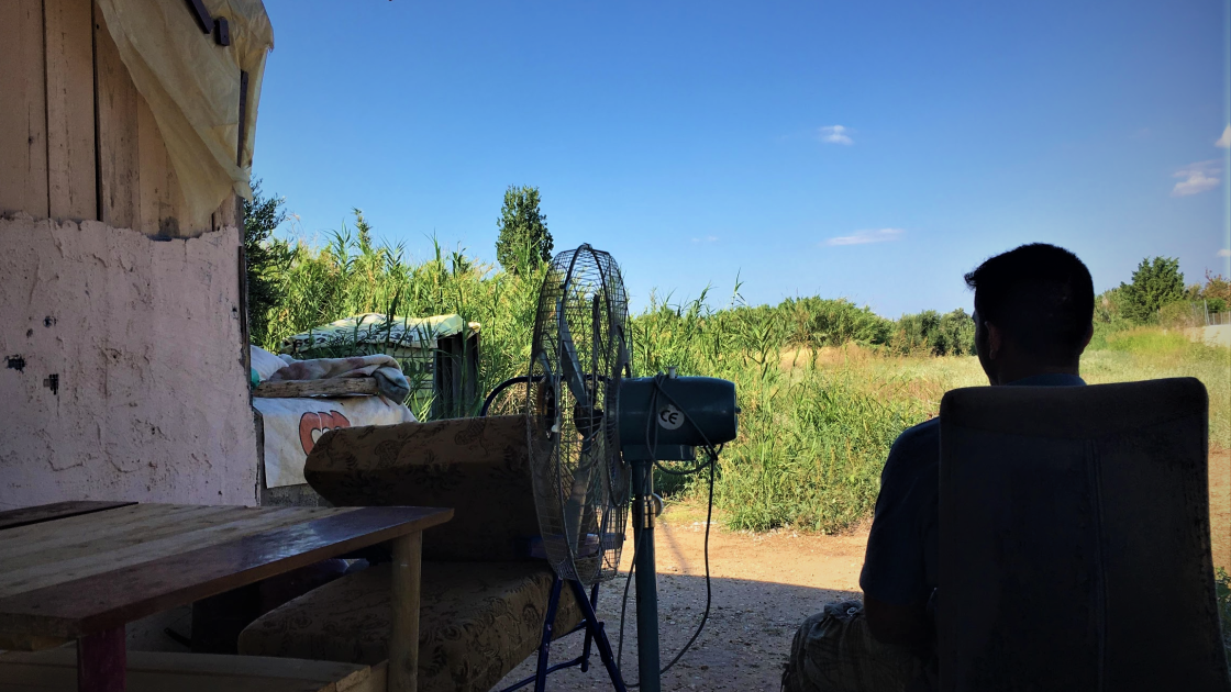 a-pakistani-teenager-in-greece.-photo-by-divya-mishra.png A boy sits on a chair outside with his back to the camera. To his left is the side of a wooden house, and next to it is a wood table, a metal chair with falling cushions, and a fan.