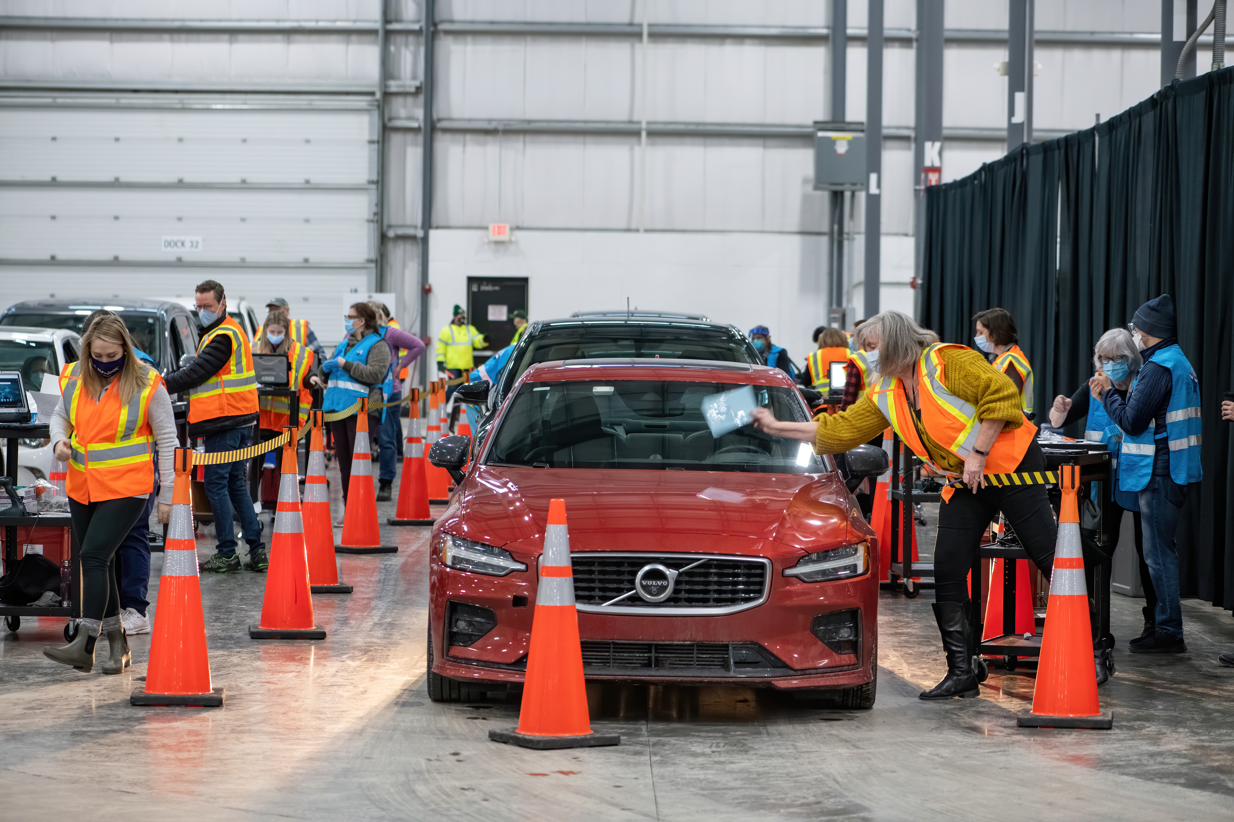 shutterstock_1908098632.jpeg Workers in masks and reflective vests distribute vaccines as people wait in cars.