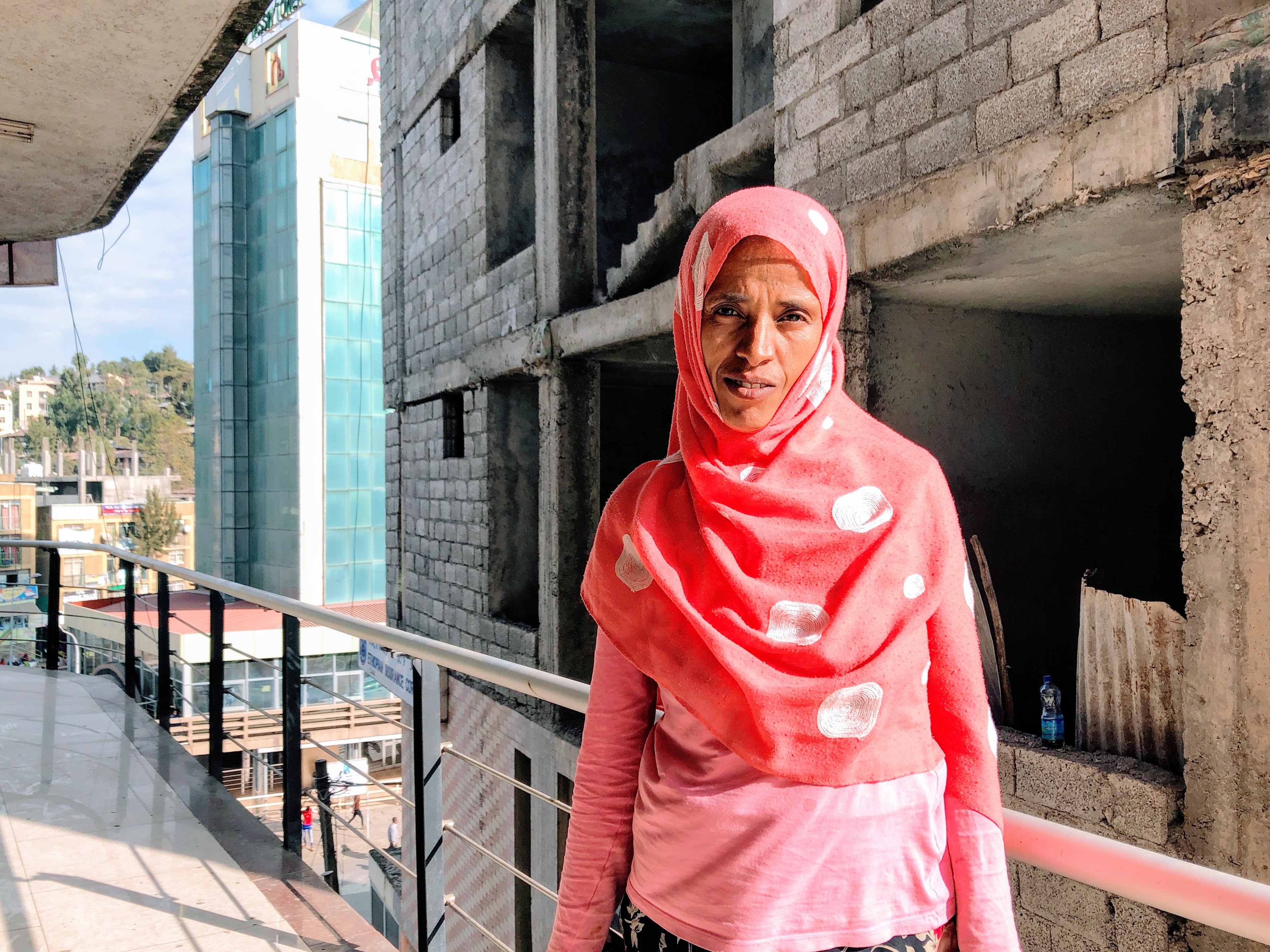 zebiba.jpg A portrait of an Ethiopian woman standing on a balcony. She is wearing a pink, long-sleeved shirt and a pink scarf with white polka dots covers her hair.