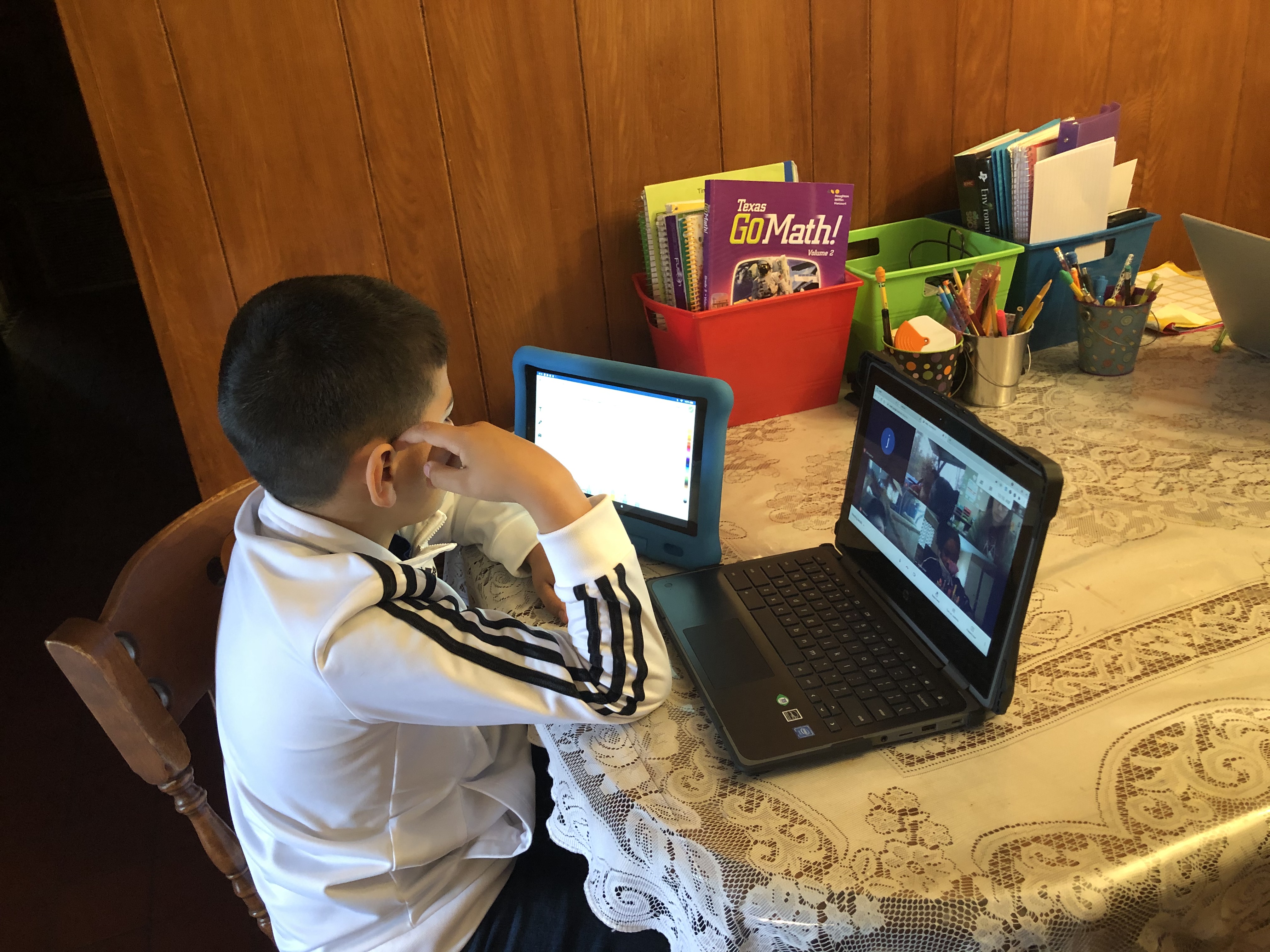 File_017.jpeg A young boy sits at a kitchen table with a tablet and laptop in front of him. On the laptop, he has a Zoom meeting open with four other attendees. On the table, there are three cups holding writing utensils and red, green, and blue containers holding notebooks and papers.