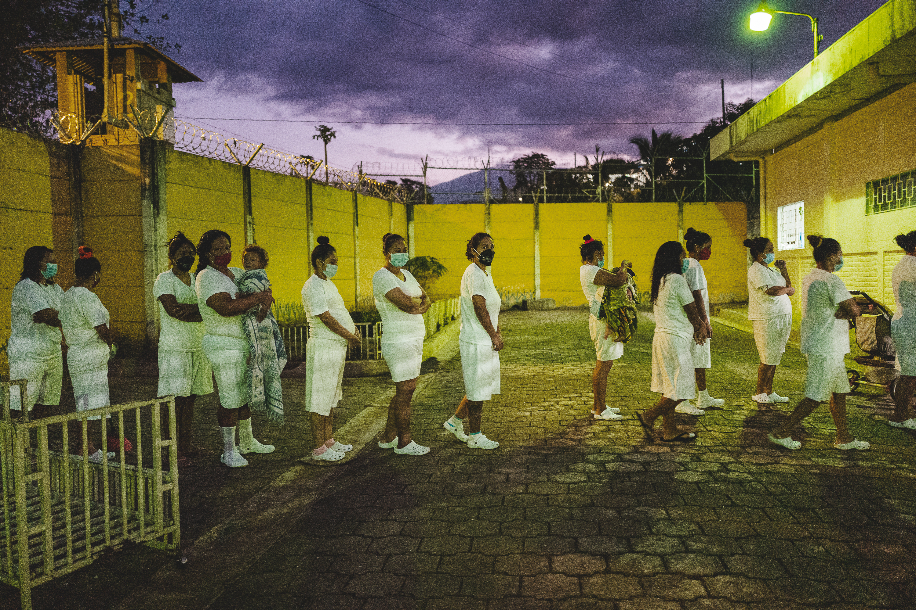 LOBA-ANAMARIA-AREVALOGOSEN-DIASETERNOS-0018.jpg Waiting to go inside, a line of women stand in a yard enclosed by a wall that is topped with barbed wire. The women wear the same all-white uniform of a t-shirt, shorts, and crocs, and they all wear masks. Two women are holding small children. On the right of the frame is a stroller.