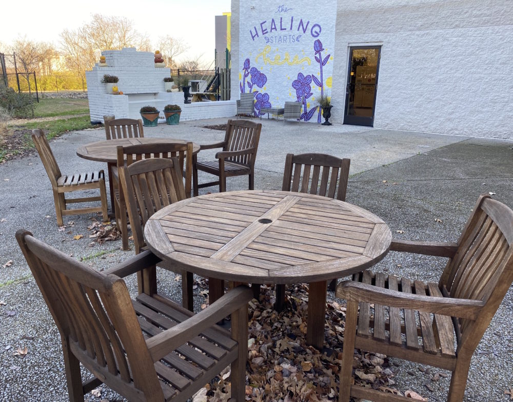 Sexual-Assault-Center_Senjanovic-1000x784.jpeg An outdoor space with sets of tables and chairs with potted plants. A mural on a wall reads, "The healing starts here."