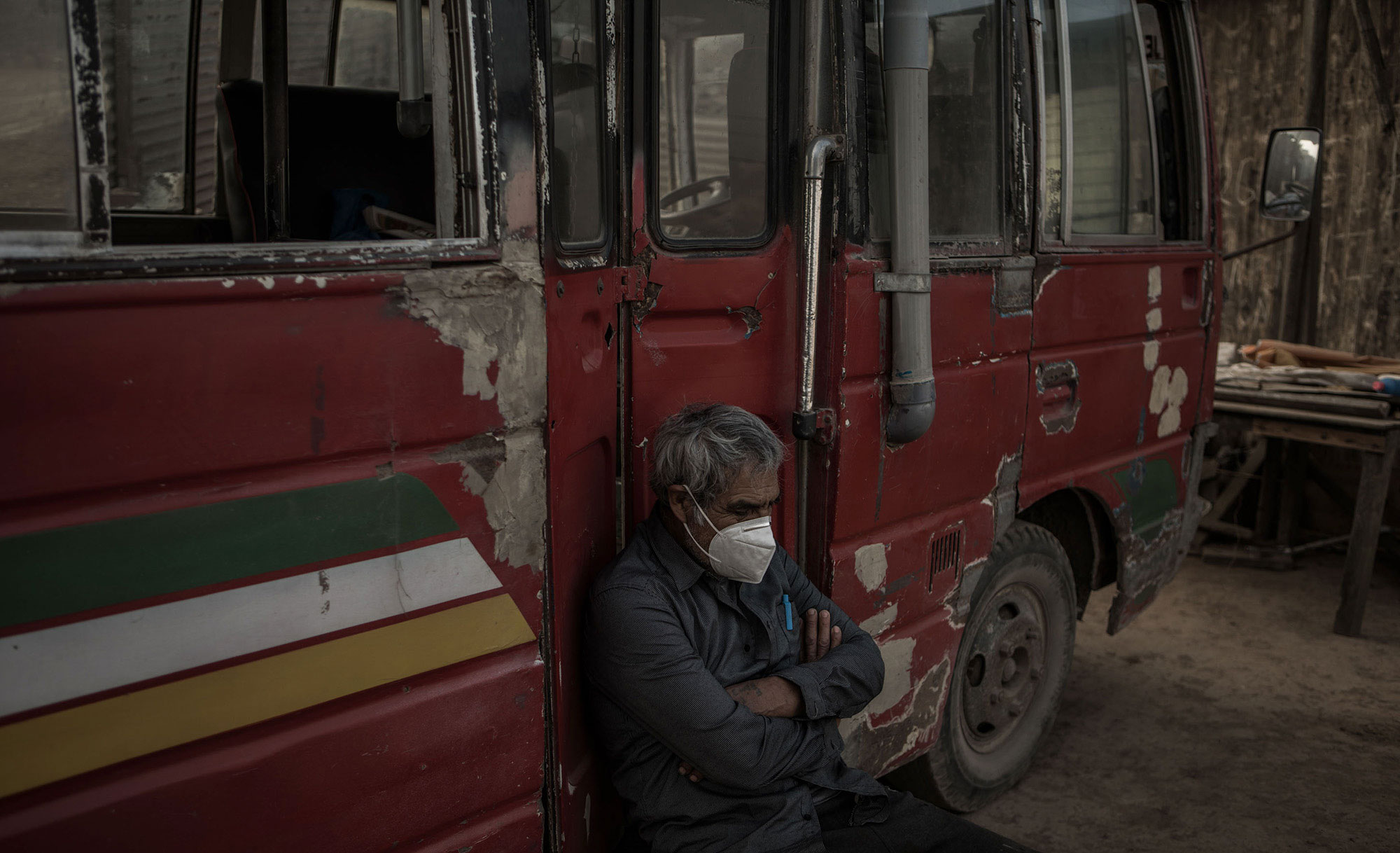 A man in a medical face mask sits and leans against a red vehicle.