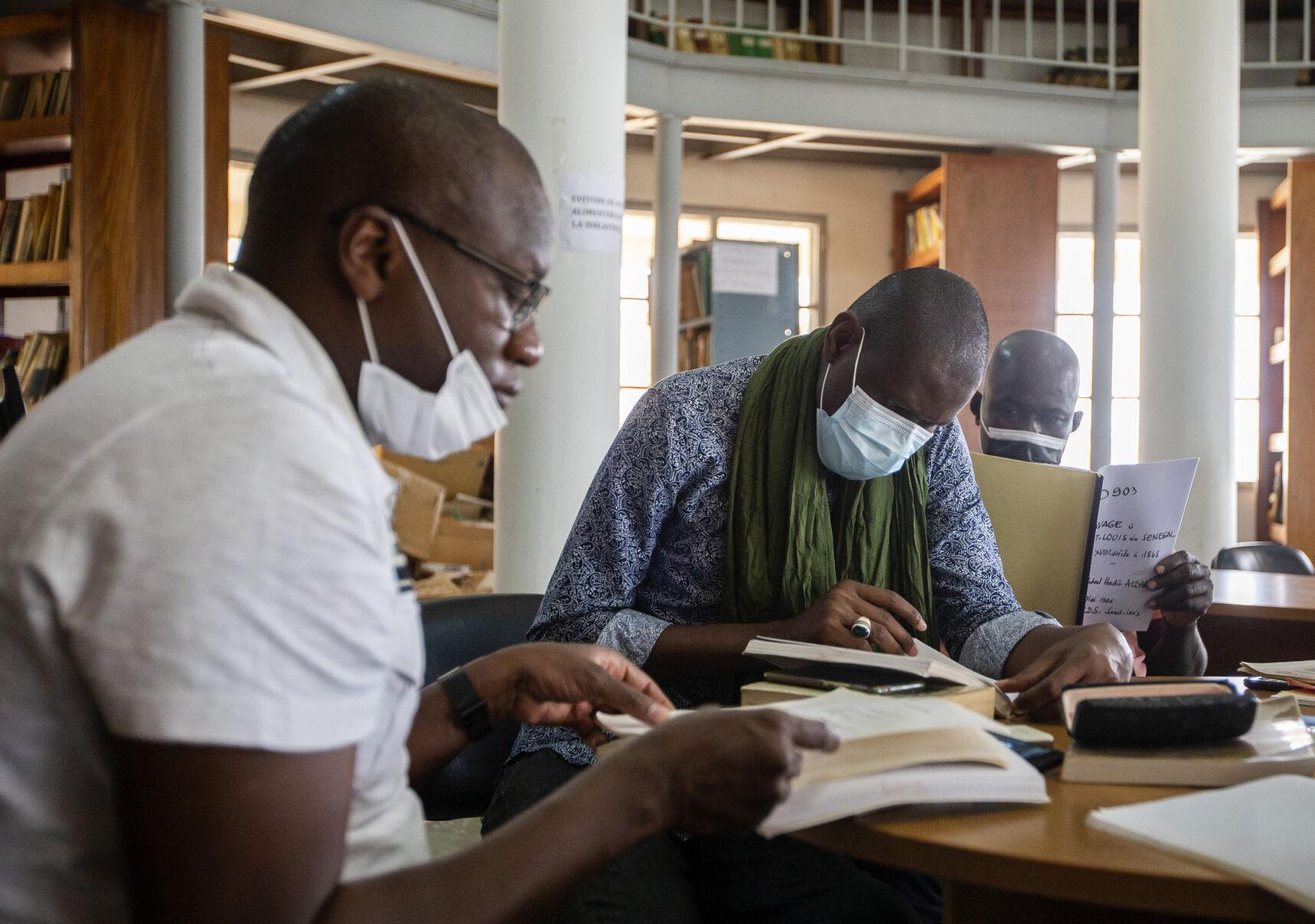 60ad2eb12ed52.image_.jpg Three people wearing medical face masks sit and read papers and books in an archive.