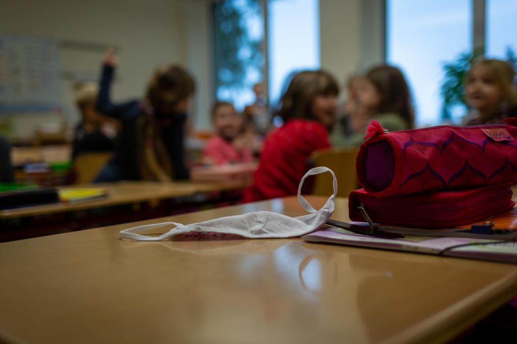 In the foreground, a white face mask sits on a school desk along with a pencil case and other supplies. In the blurred background, a group of young schoolchildren sit and talk together in a classroom.