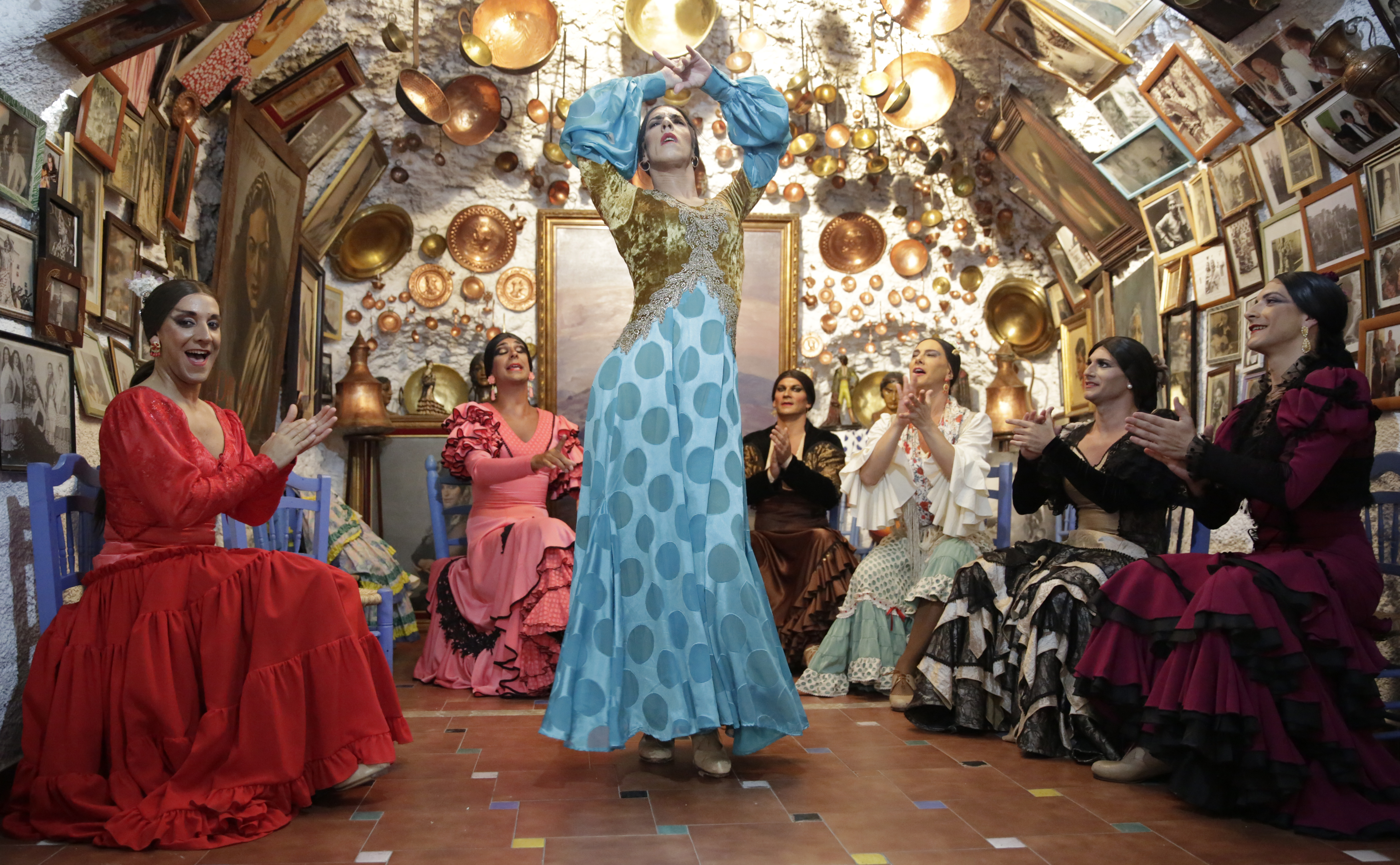 A man in drag wearing a blue and gold dress dances in the middle of a room. Sitting around the perimeter of the room and clapping along are five other men in drag. The walls are covered in gold decorations and framed pictures.