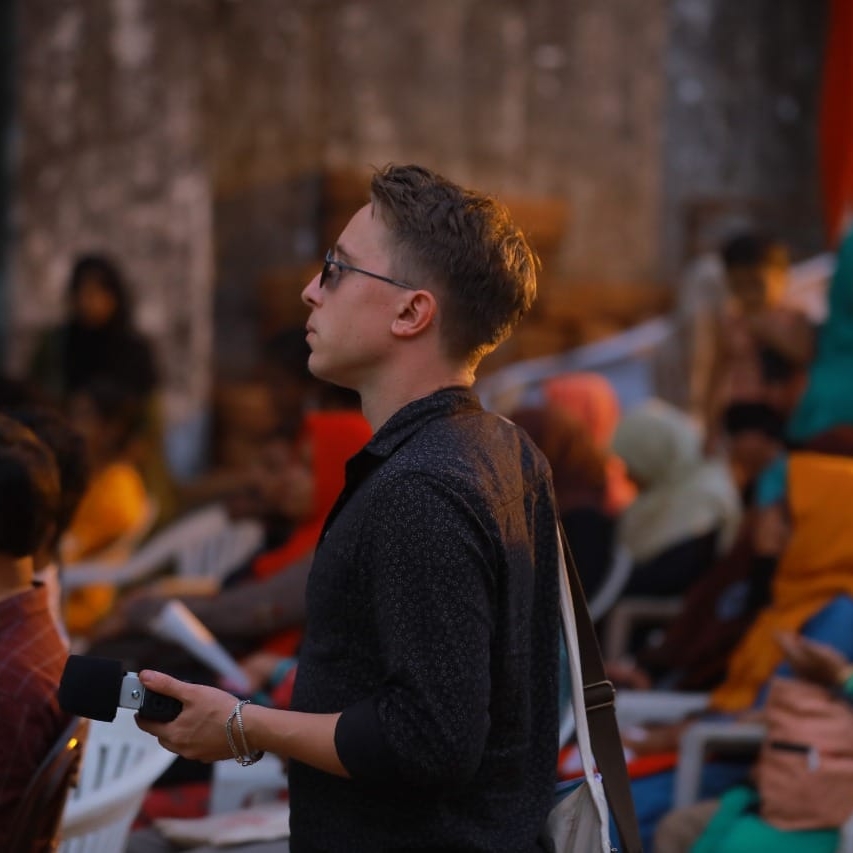 Côme Bastin A man wears a black shirt and sunglasses. He carries a microphone. In the foreground, there are people sitting on plastic chairs.