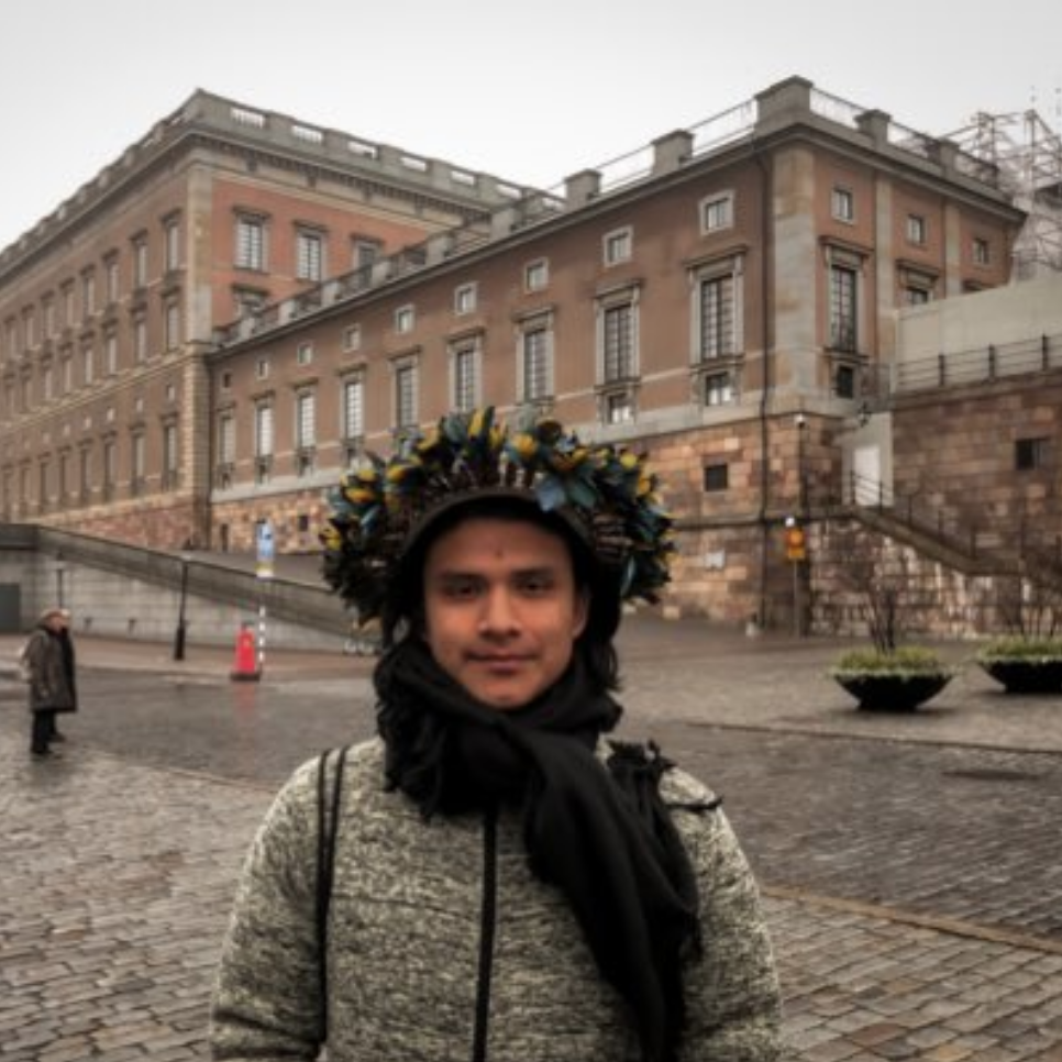 A person wears a grey winter jacket and an Indigenous headdress. In the foreground is a building.