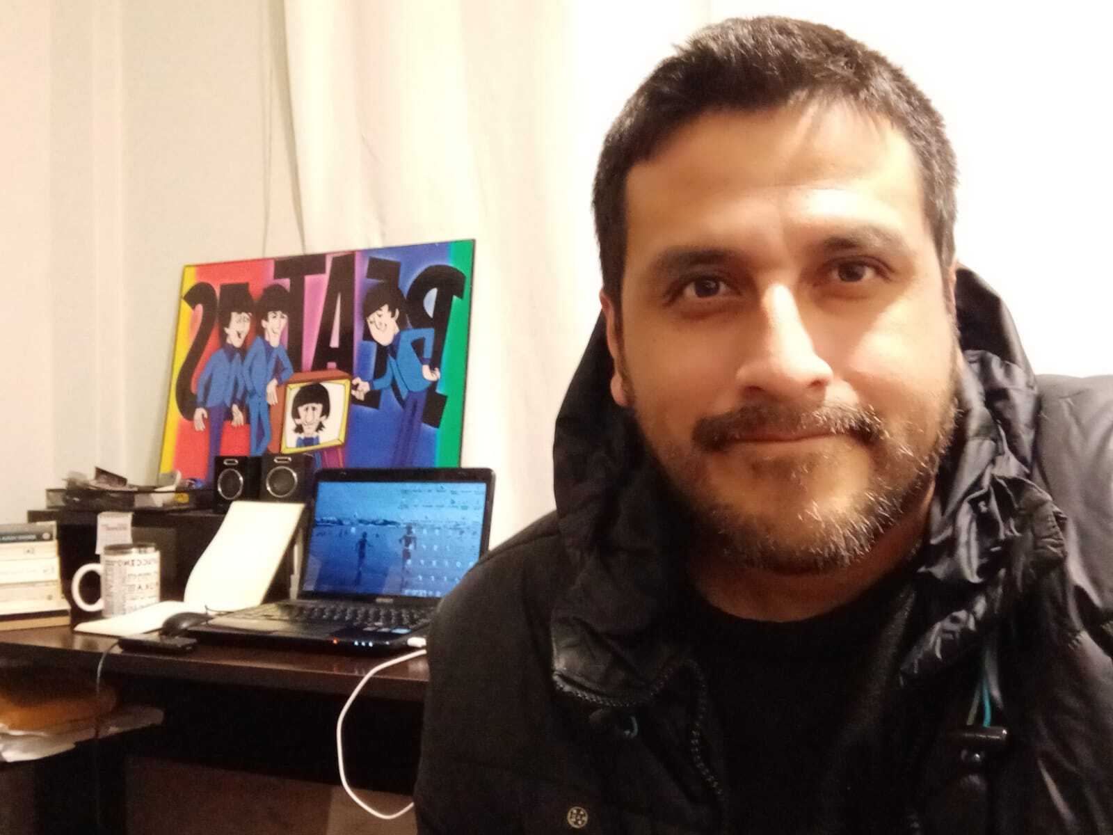 A person wears a black jacket. In the foreground, there is a study desk with a laptop and a poster board.