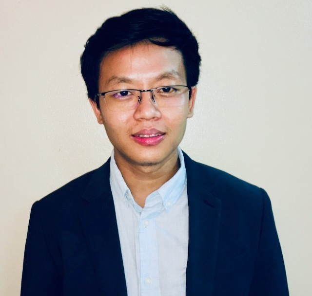 A person with a black jacket, light blue shirt, and glasses.