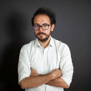 A man with short brown hair, black eyeglasses, a white shirt, and he crosses his arms.