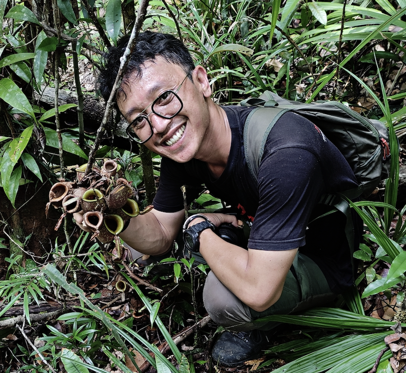 A man wears a black shirt and trekking pants. He has eyeglasses and is holding a plant.