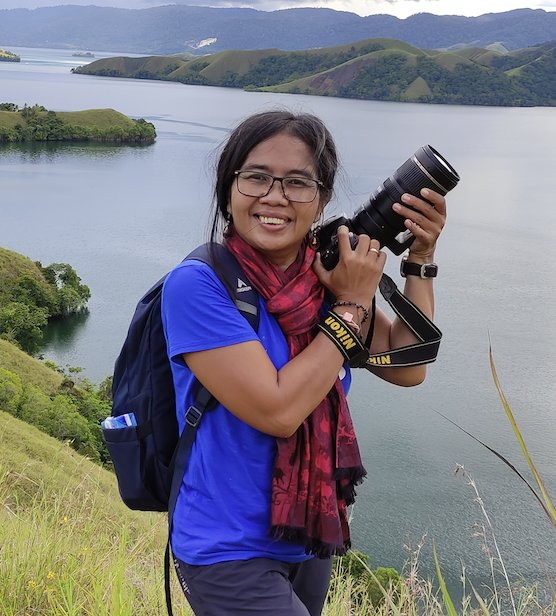 A woman holds a camera in her hands. She wears glasses, a purple shirt and jeans.