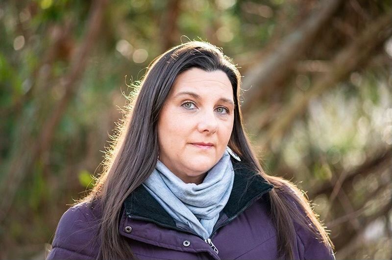 Jennifer Randolph A woman wears a scarf and purple coat in front of trees