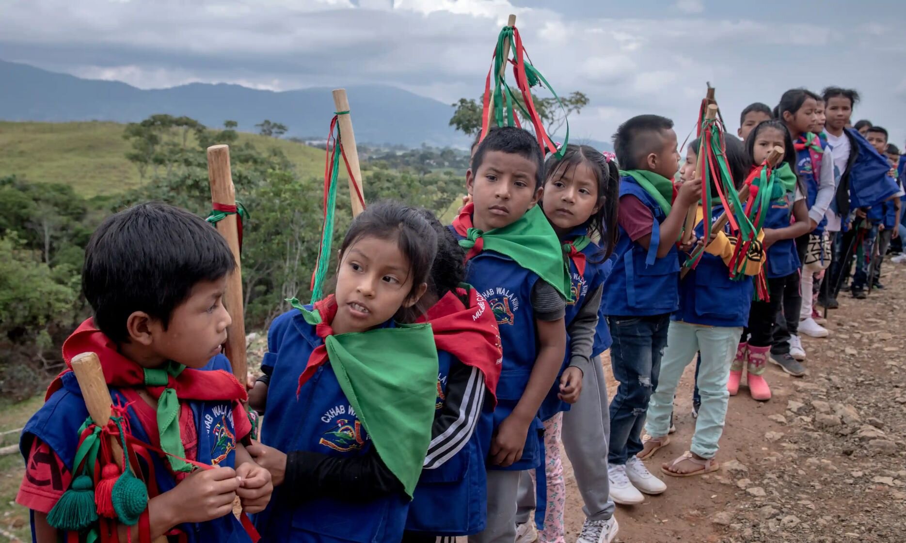 'We must not show fear' Children in blue vests and green-and-red bandanas tied around their necks line up on a mountain trail. Each carries a wood staff.
